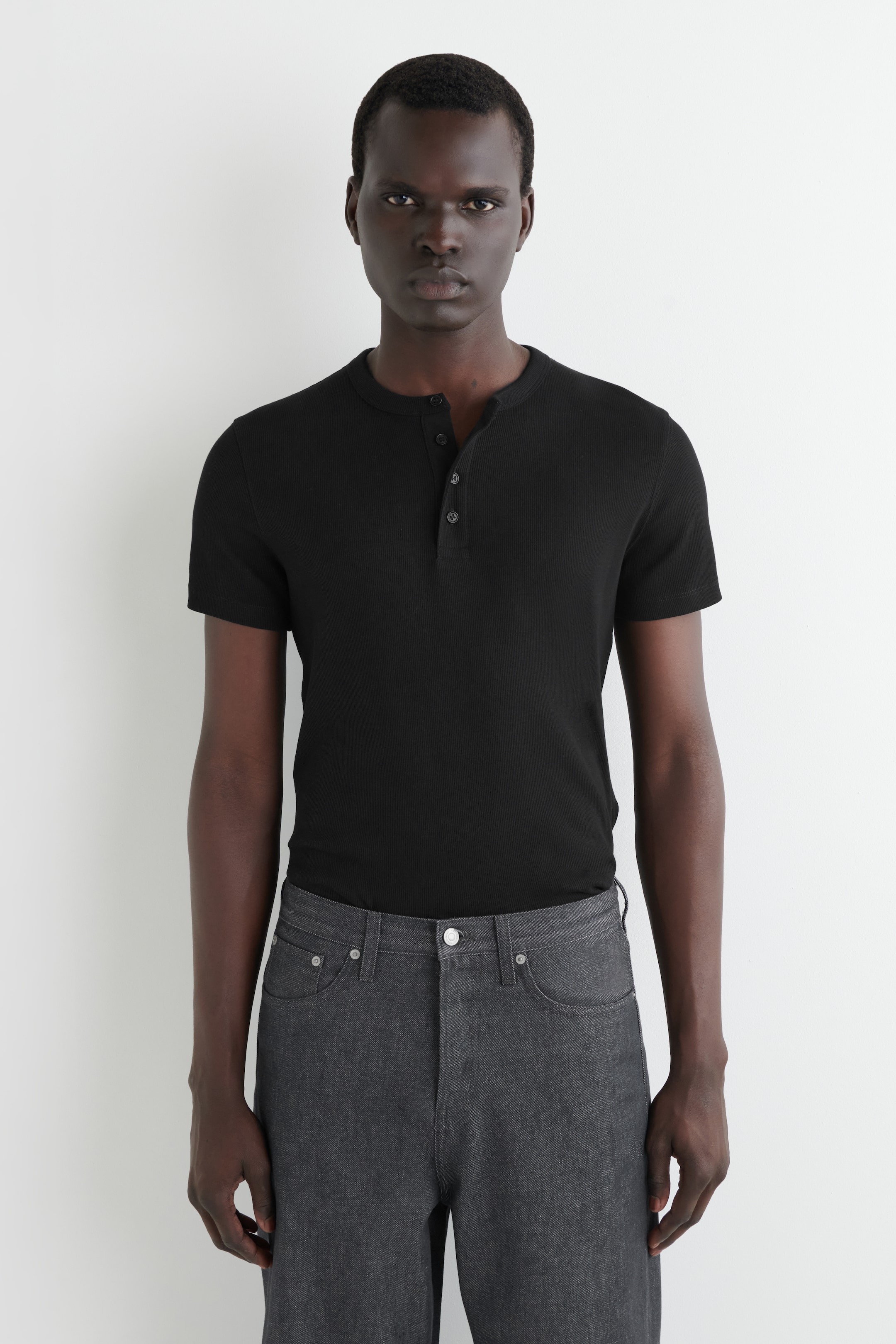 Agrandir l'image: Un t-shirt Henley noir à manches courtes avec une texture en maille côtelée et une patte de boutonnage à trois boutons est présenté. Il est porté rentré dans un jean gris foncé à coupe décontractée avec un style à cinq poches visible.