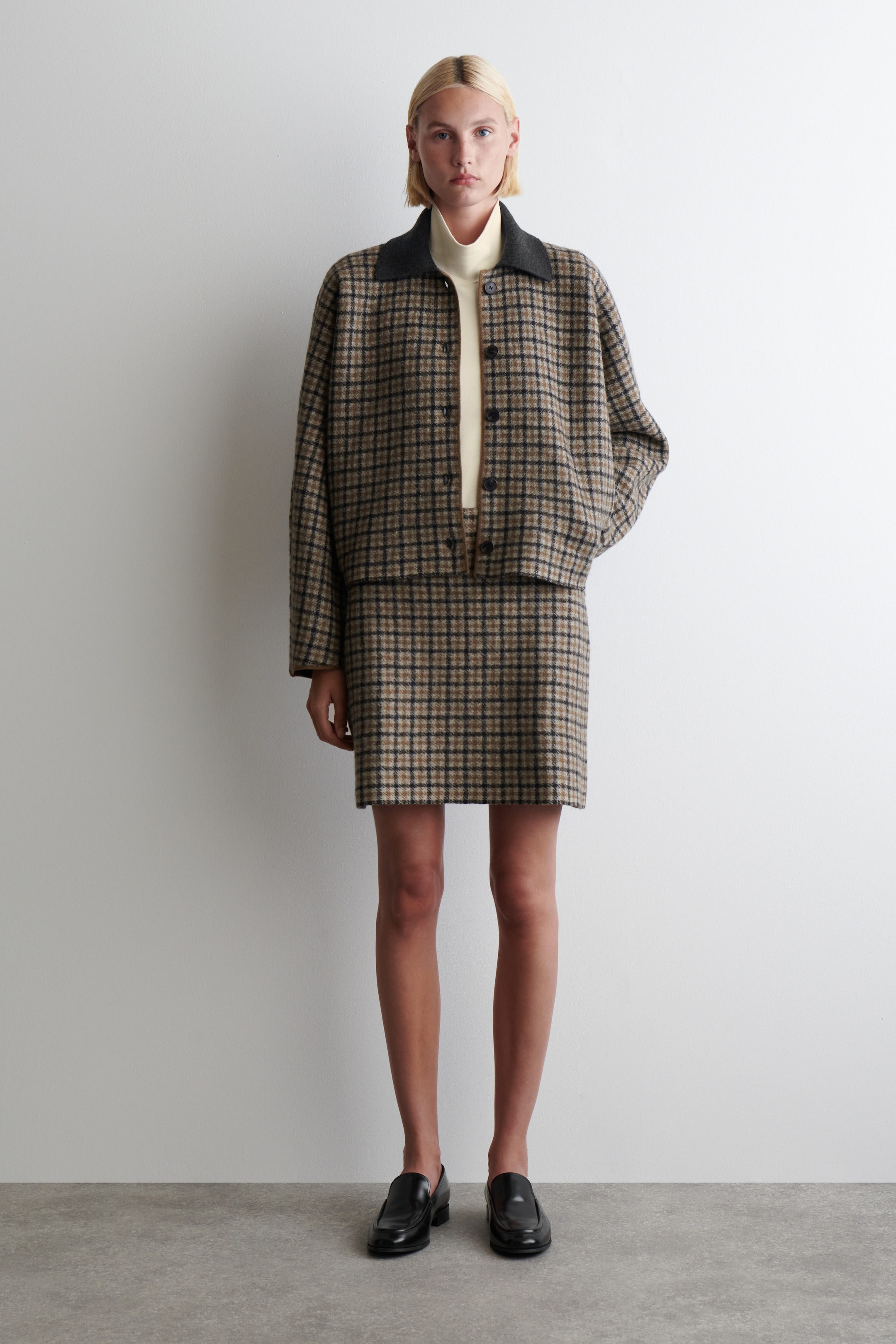 Grösseres Bild ansehen: Ein zweiteiliges Set in Braun, Schwarz und Beige mit Karomuster wird präsentiert, bestehend aus einer kurzen Jacke und einem Minirock. Die Jacke verfügt über einen dunkel kontrastierenden spitzen Kragen und einen vorderen Knopfverschluss und wird über einem hellen Rollkragenpullover getragen; der passende karierte Minirock zeigt eine gerade Silhouette.