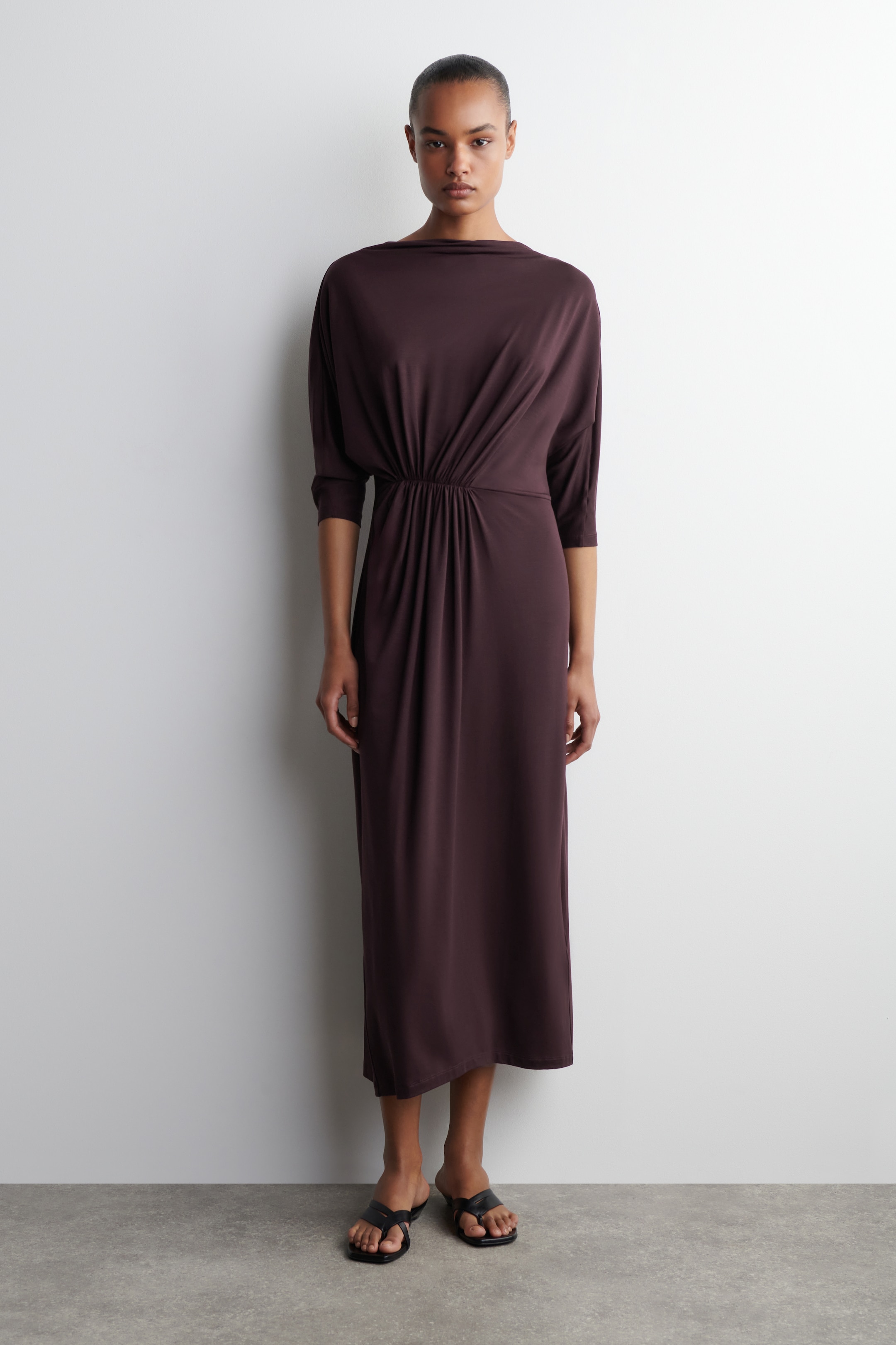 Größeres Bild anzeigen: Ein dunkel pflaumenbraunes Midikleid wird von vorne gezeigt, mit einem Bootsausschnitt und dreiviertellangen Dolman-Ärmeln. Das Kleid ist in der Taille mit gerafften Details tailliert, was ein drapiertes Oberteil und einen fließenden Rock schafft.