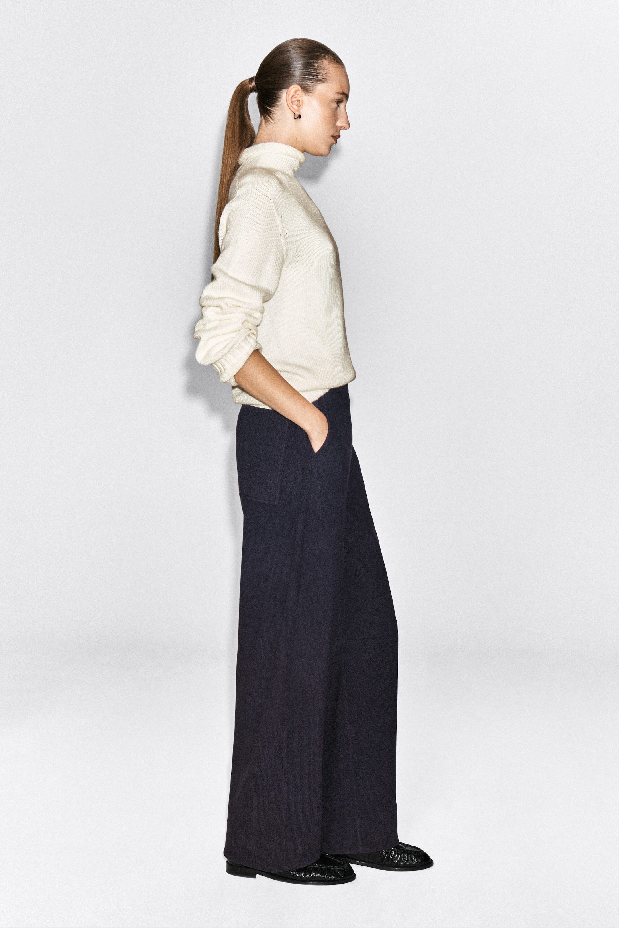 The model is wearing dark navy corduroy-jersey wide-leg pants, standing in profile with one hand resting in the side pocket. The pants are styled with a cream-colored high-neck knit sweater and black shoes.