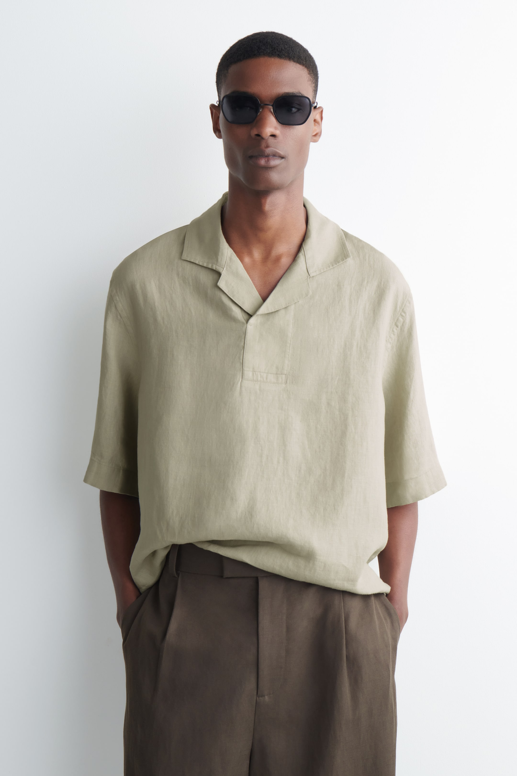 Agrandir l'image: Une chemise en lin vert kaki clair à manches courtes est portée, avec un col camp et une patte de boutonnage partielle sans boutons. Elle offre une coupe décontractée, tombant non rentré sur un pantalon foncé.