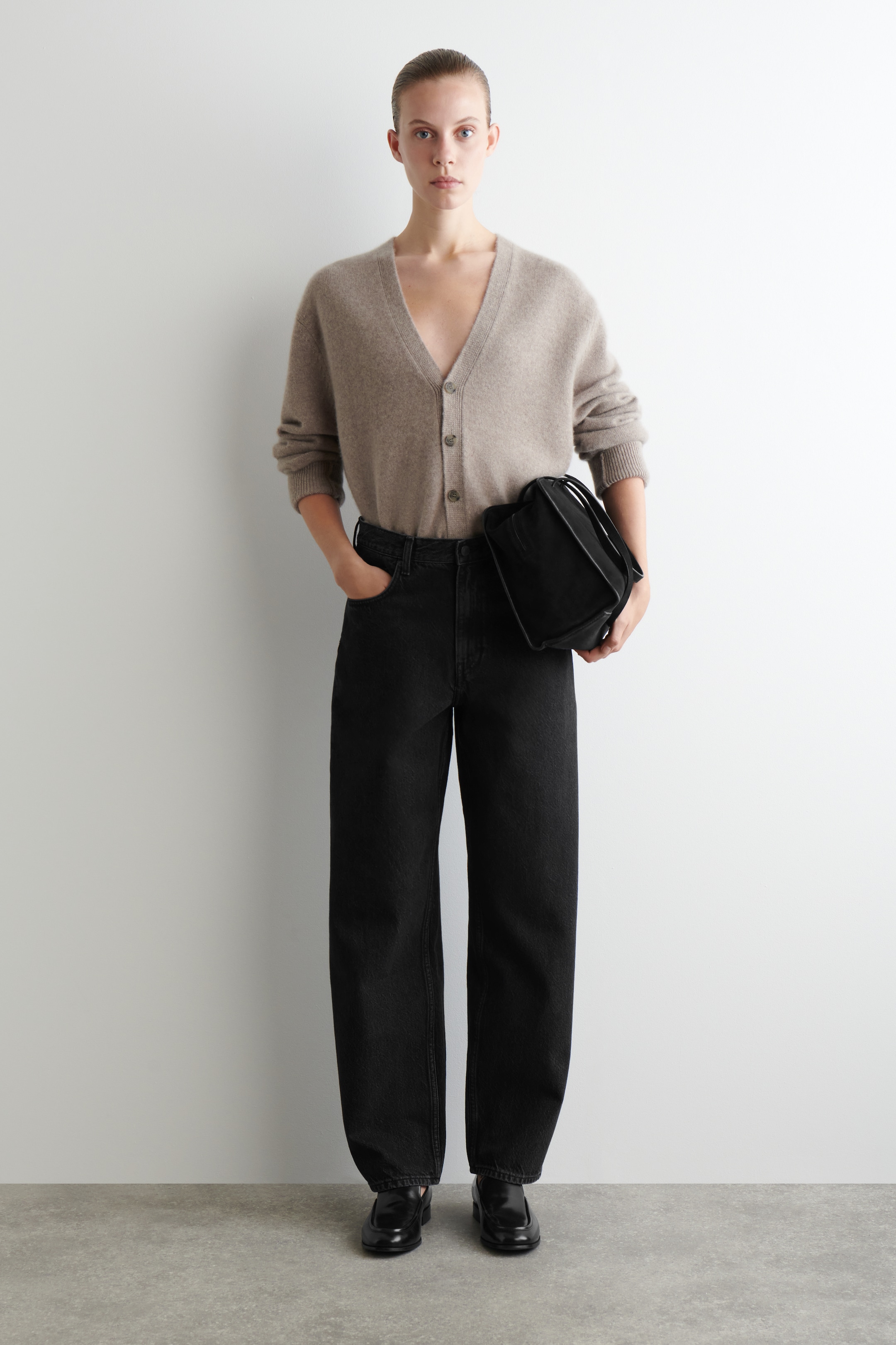 Visualizza immagine più grande: Un cardigan marrone chiaro con scollo a V, chiusura frontale con bottoni e polsini a coste viene presentato da un'angolazione frontale, abbinato a jeans neri a gamba larga. Una borsa nera strutturata è tenuta in una mano e mocassini neri sono indossati ai piedi.
