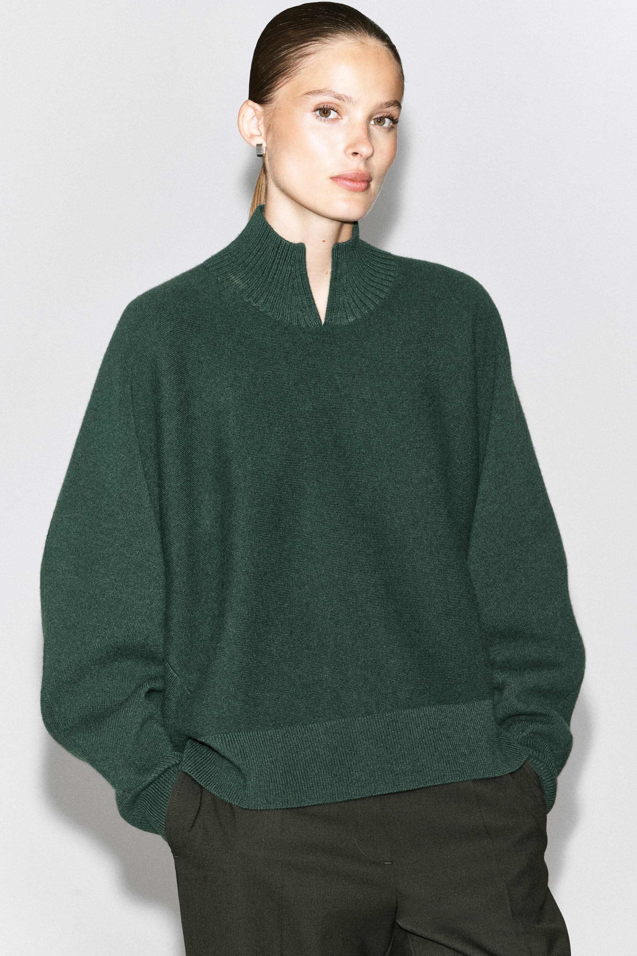 Un chandail en laine tricotée vert foncé est porté par le modèle, avec une silhouette décontractée et un col montant côtelé avec détail en V à l’avant. Le chandail est agencé avec un pantalon foncé, tandis que le modèle a les mains dans les poches. Un chandail en laine tricotée vert foncé est porté par le modèle, avec une silhouette décontractée et un col montant côtelé avec détail en V à l’avant. Le chandail est agencé avec un pantalon foncé, tandis que le modèle a les mains dans les poches.