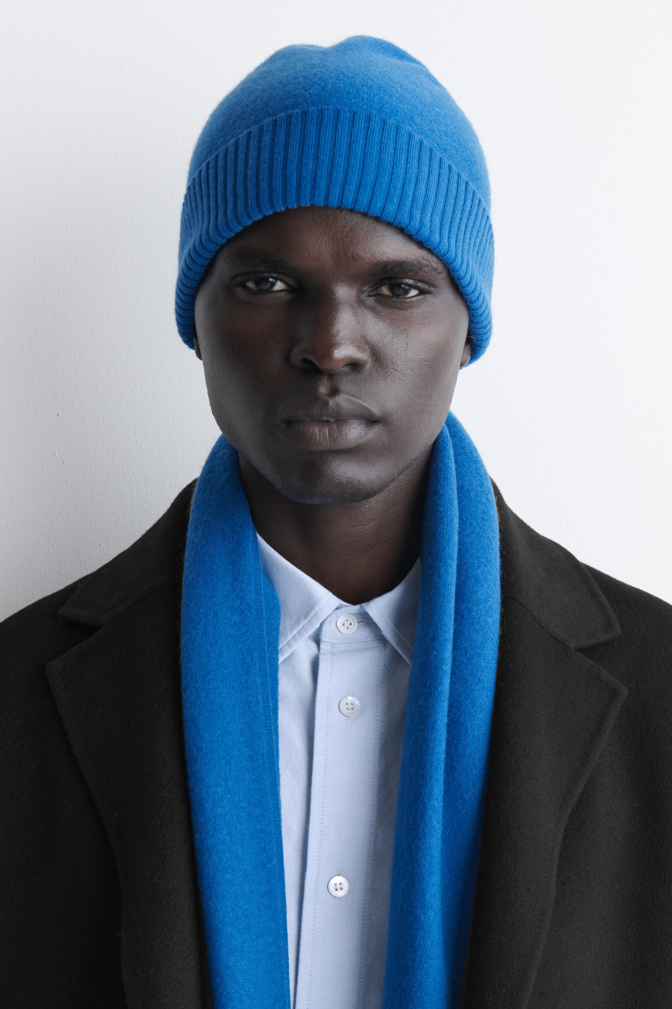 Zobacz większy obraz: Jasnoniebieska dzianinowa czapka typu beanie z fakturowanym, wełnianym korpusem i prążkowanym wywijanym brzegiem jest noszona przez modela ustawionego przodem do aparatu. Beanie zestawiono z dopasowanym niebieskim szalikiem swobodnie ułożonym wokół szyi, na czarnym płaszczu i jasnoniebieskiej koszuli zapinanej na guziki.
