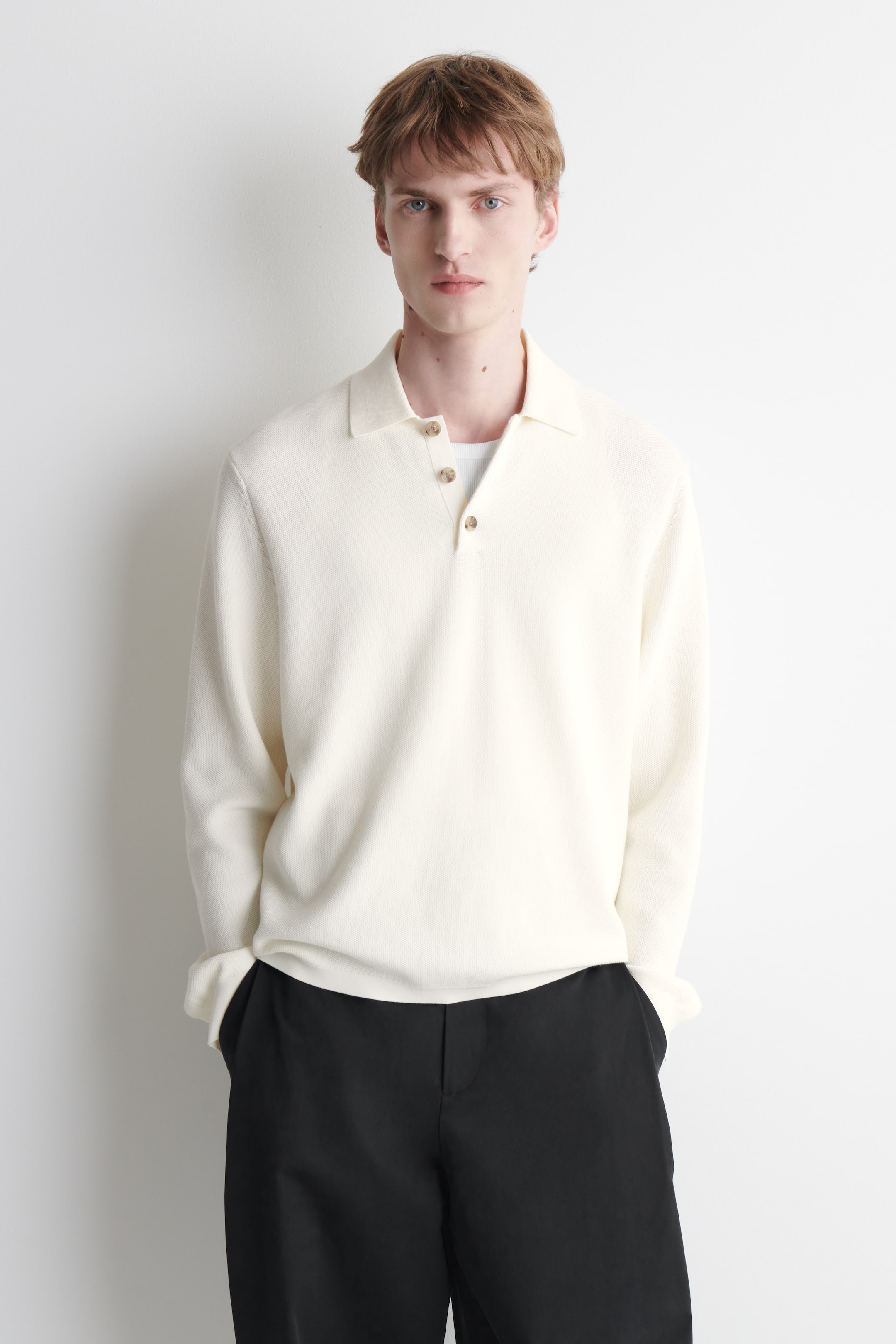 Größeres Bild ansehen: Ein cremefarbenes Langarm-Poloshirt wird getragen und zeichnet sich durch eine strukturierte Strickoptik, einen breiten Kragen sowie eine Drei-Knopf-Leiste mit hellbraunen Knöpfen aus; darunter ist durch die offene Knopfleiste ein weißes Unterhemd sichtbar, und die langen Ärmel sind hochgekrempelt.