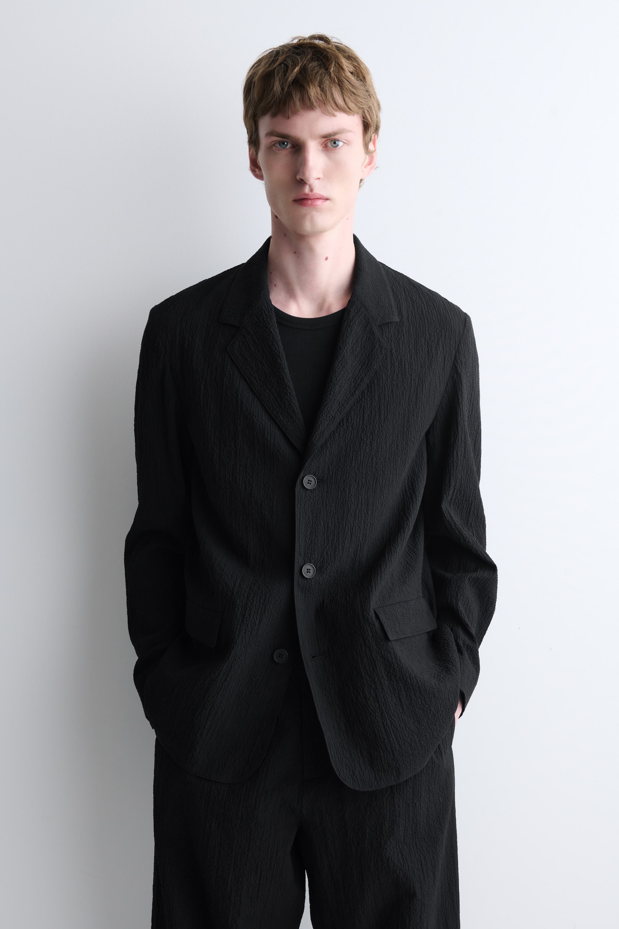 Agrandir l'image: Un blazer texturé noir et un pantalon assorti sont présentés, face à l'avant. Le blazer est doté de revers crantés, d'une fermeture à deux boutons et de deux poches plaquées, porté sur un t-shirt noir à col rond.