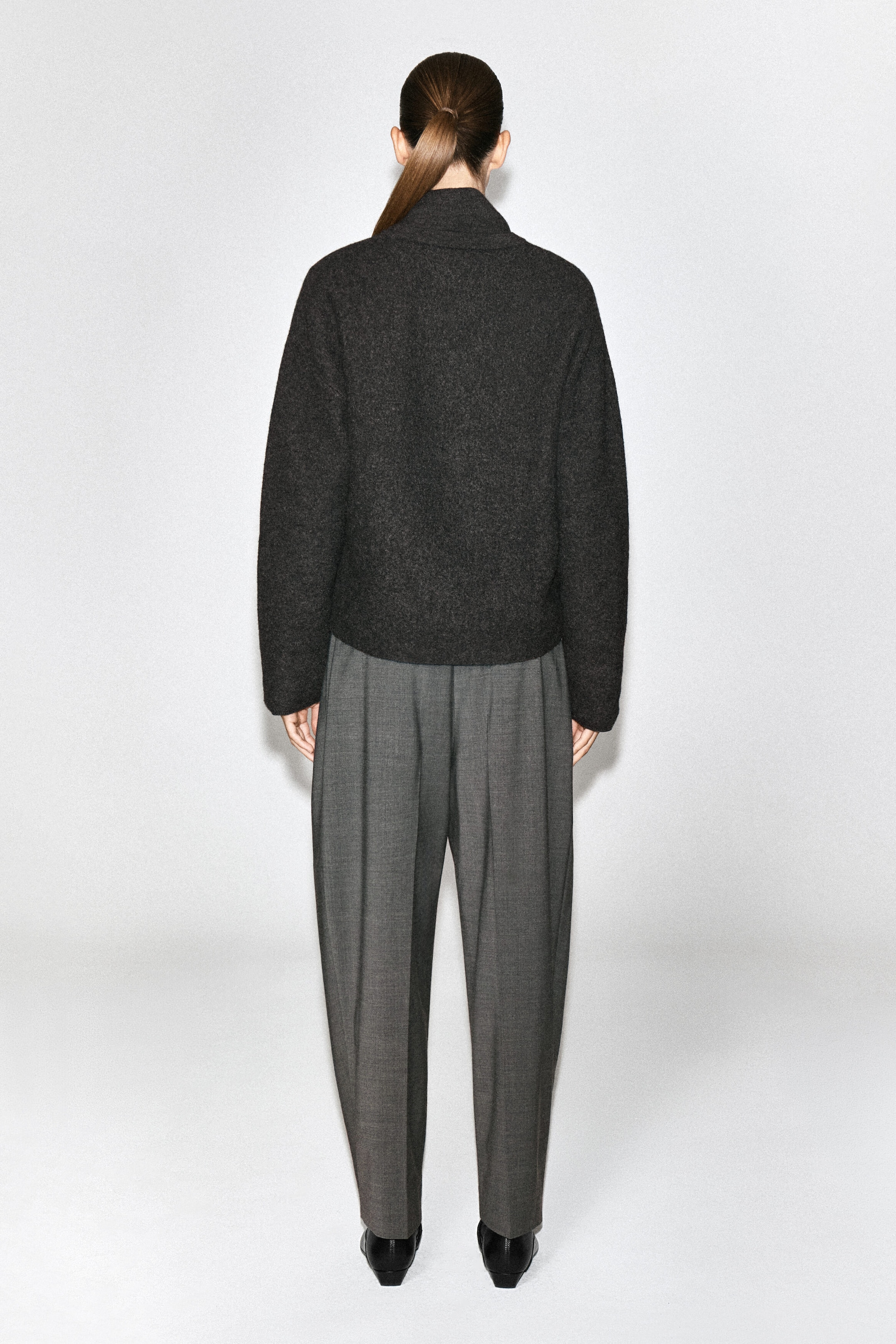 A dark gray boiled-wool sweater featuring a high, layered collar and long sleeves is being worn by the model, shown from the back. The sweater is paired with gray pleated trousers.