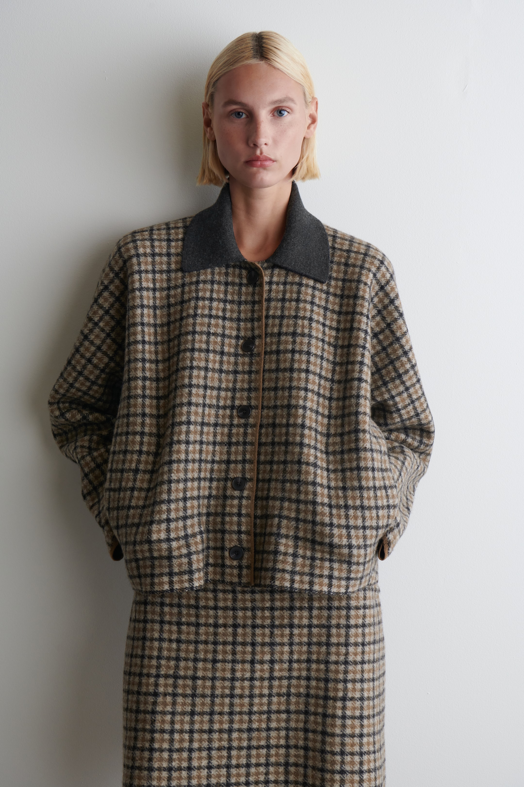 View larger image: A front-facing jacket and a matching skirt are being shown. The jacket is featuring a brown and black plaid pattern, a contrasting dark grey collar, a button-front closure, and side pockets where the wearer's hands are resting. The coordinated skirt is displaying the same plaid pattern, completing the set.