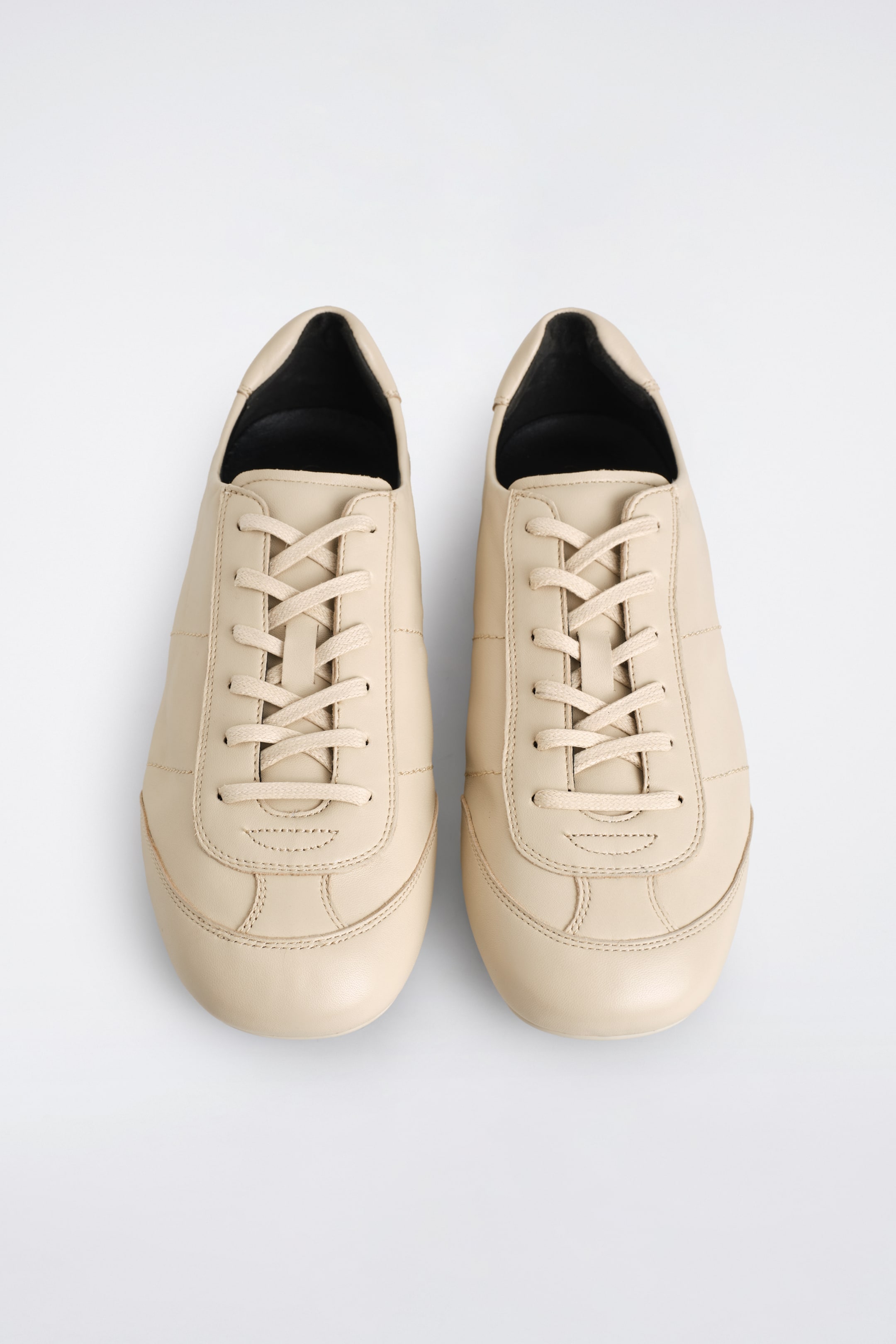 Größeres Bild ansehen: Ein Paar hellbeige Ledersneaker wird aus der VOGELPERSPEKTIVE gezeigt, mit stromlinienförmigen Obermaterialien und tonalen Ziernähten, passenden Schnürverschlüssen und einem schwarzen Innenfutter.