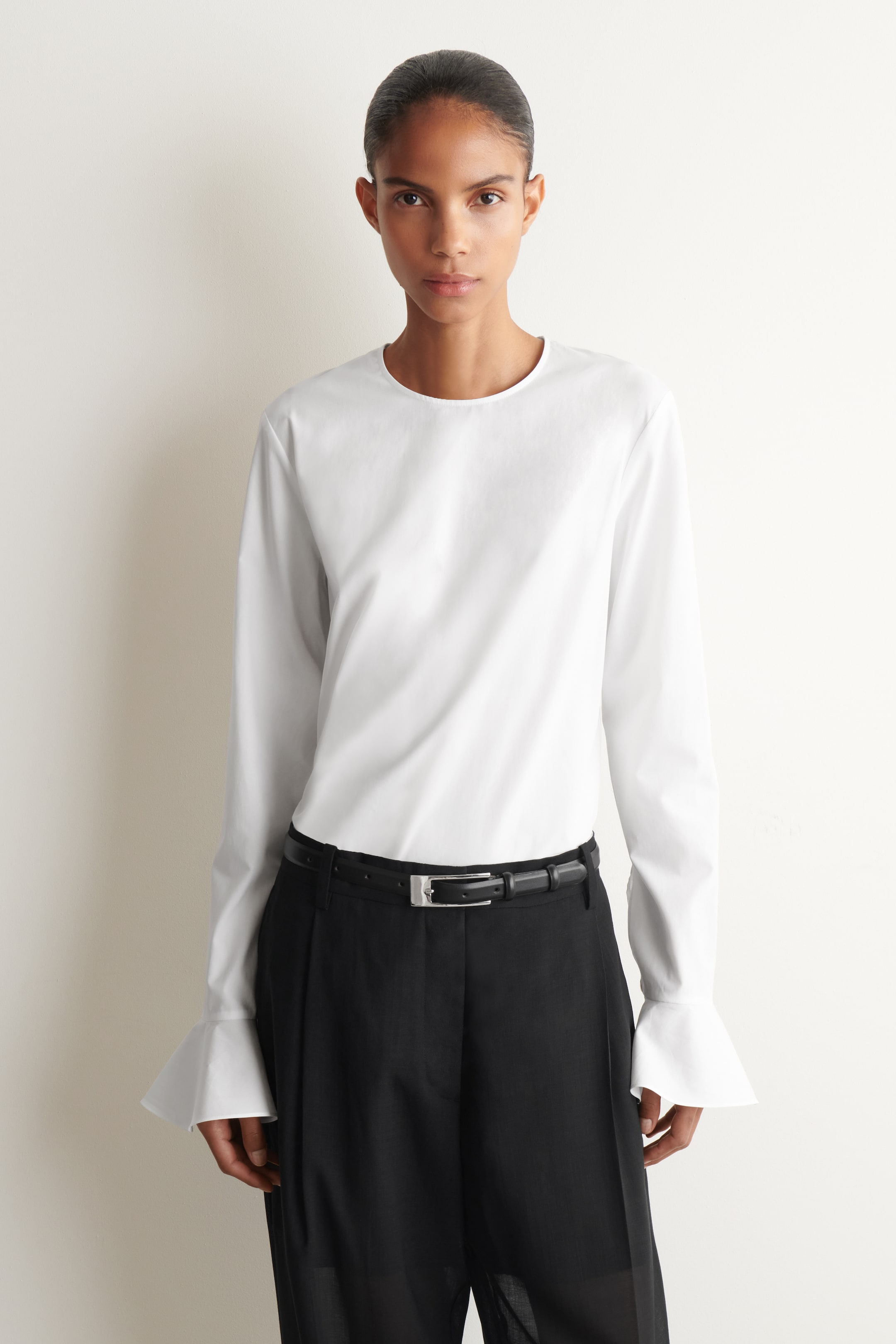 Ver imagen más grande: Una blusa blanca de manga larga presenta cuello redondo y puños acampanados. Está metida por dentro de un pantalón negro de pernera ancha y pinzas, ajustado a la cintura por un fino cinturón de cuero negro con hebilla de tono plateado.