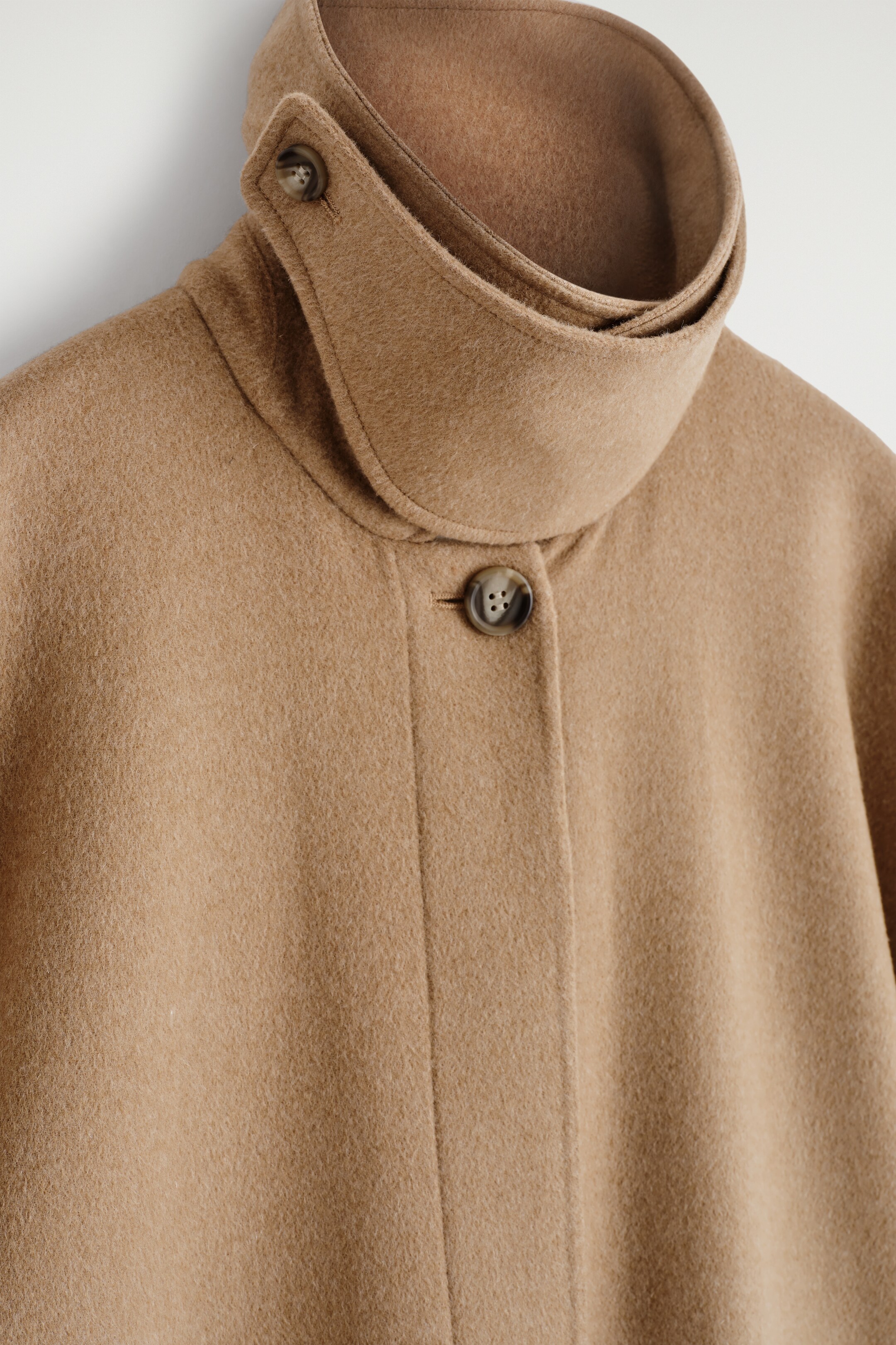 Agrandir l'image: Un long manteau en laine double face est présenté de face, avec un col montant en couches. Une patte à bouton sécurise le col, et un seul bouton ferme la patte de boutonnage avant.
