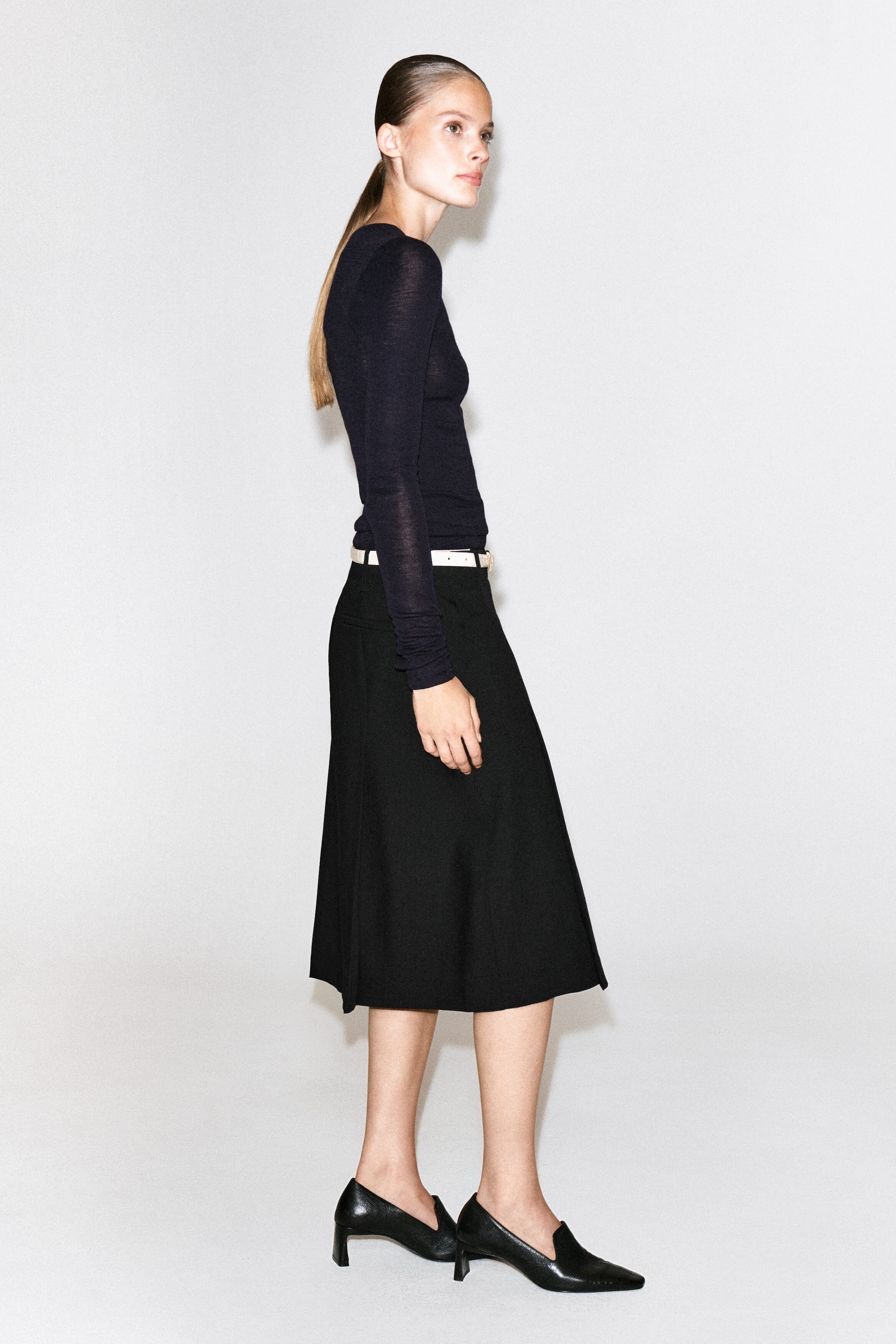 The model is wearing a black wool midi skirt with box pleats and a structured A-line silhouette. It is styled with a slim white belt at the waist, a long-sleeved dark top, and black pointed-toe low-heeled shoes. The model is wearing a black wool midi skirt with box pleats and a structured A-line silhouette. It is styled with a slim white belt at the waist, a long-sleeved dark top, and black pointed-toe low-heeled shoes.