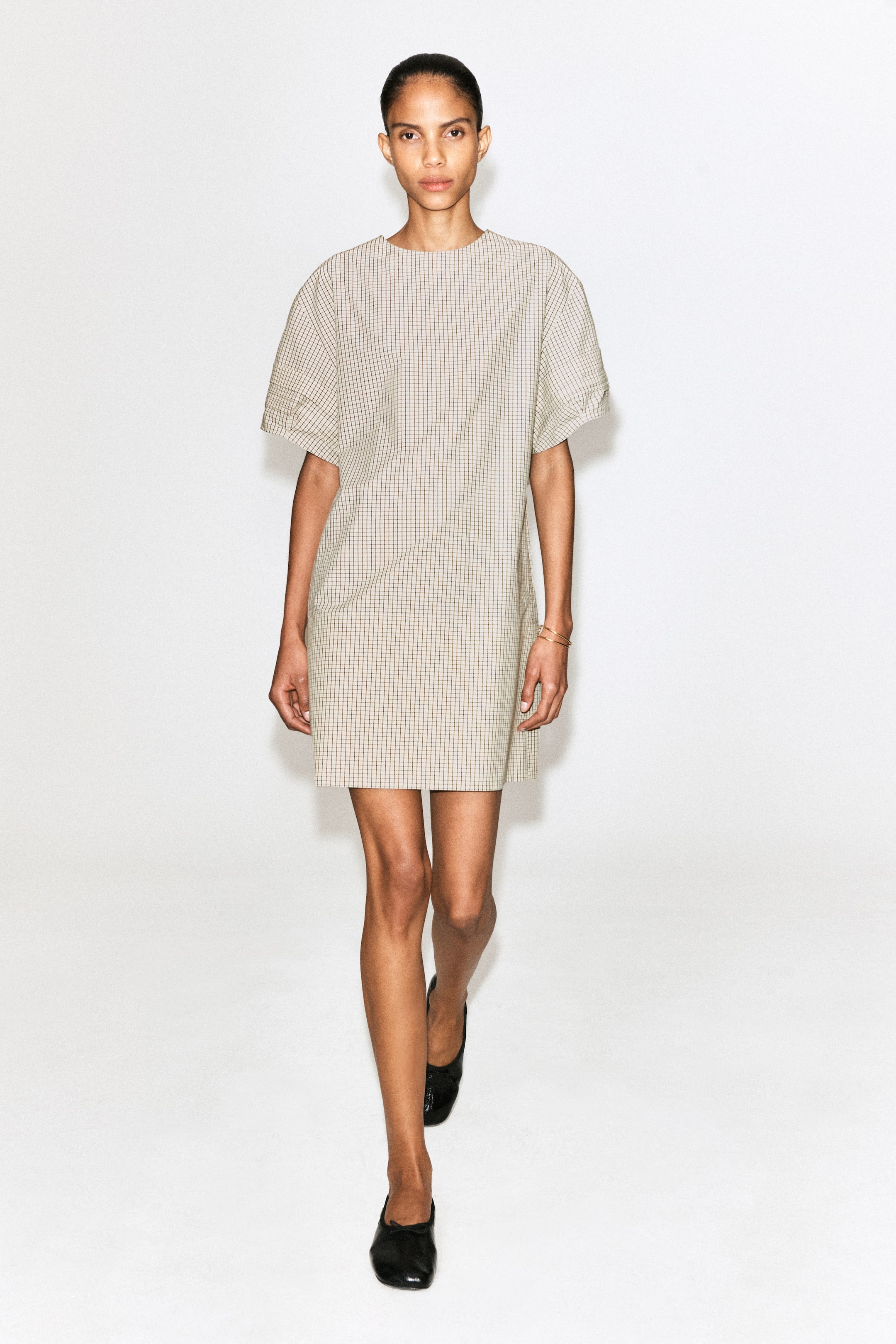 View larger image: A light-coloured mini dress with a small grid pattern is being presented from the front, featuring a rounded neckline, wide short sleeves with pintuck detailing at the cuffs, and discreet side pockets. The dress is showcasing a loose, A-line fit, falling above the knees.
