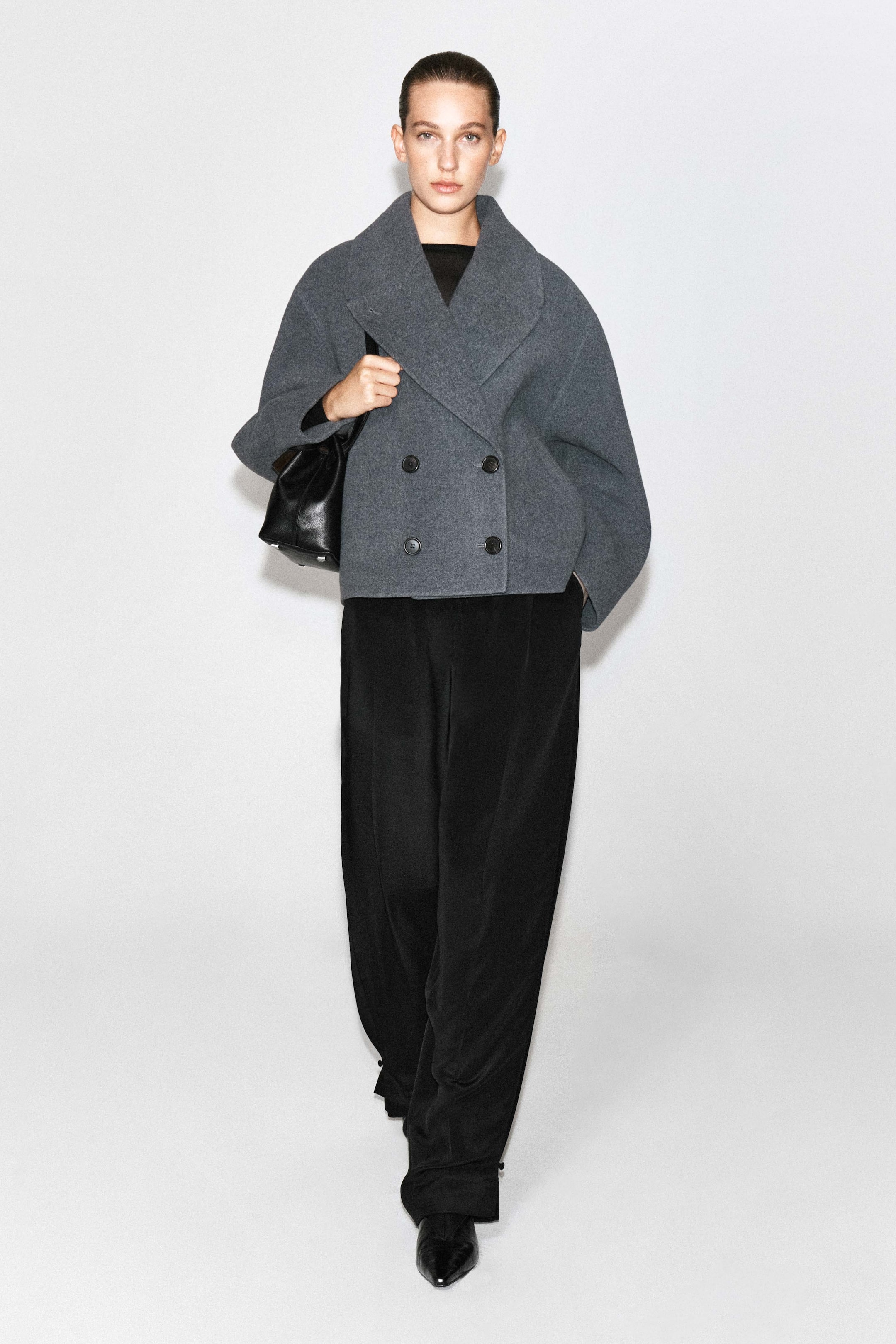 Προβολή μεγαλύτερης εικόνας: Ένα oversized διπλής όψης μάλλινο σακάκι σε γκρι απόχρωση φοριέται από το μοντέλο, με φαρδύ γιακά, κοντό μήκος και διπλό κλείσιμο με κουμπιά. Το προϊόν συνδυάζεται με φαρδύ μαύρο παντελόνι και μια μαύρη δερμάτινη τσάντα φορεμένη στον ώμο.