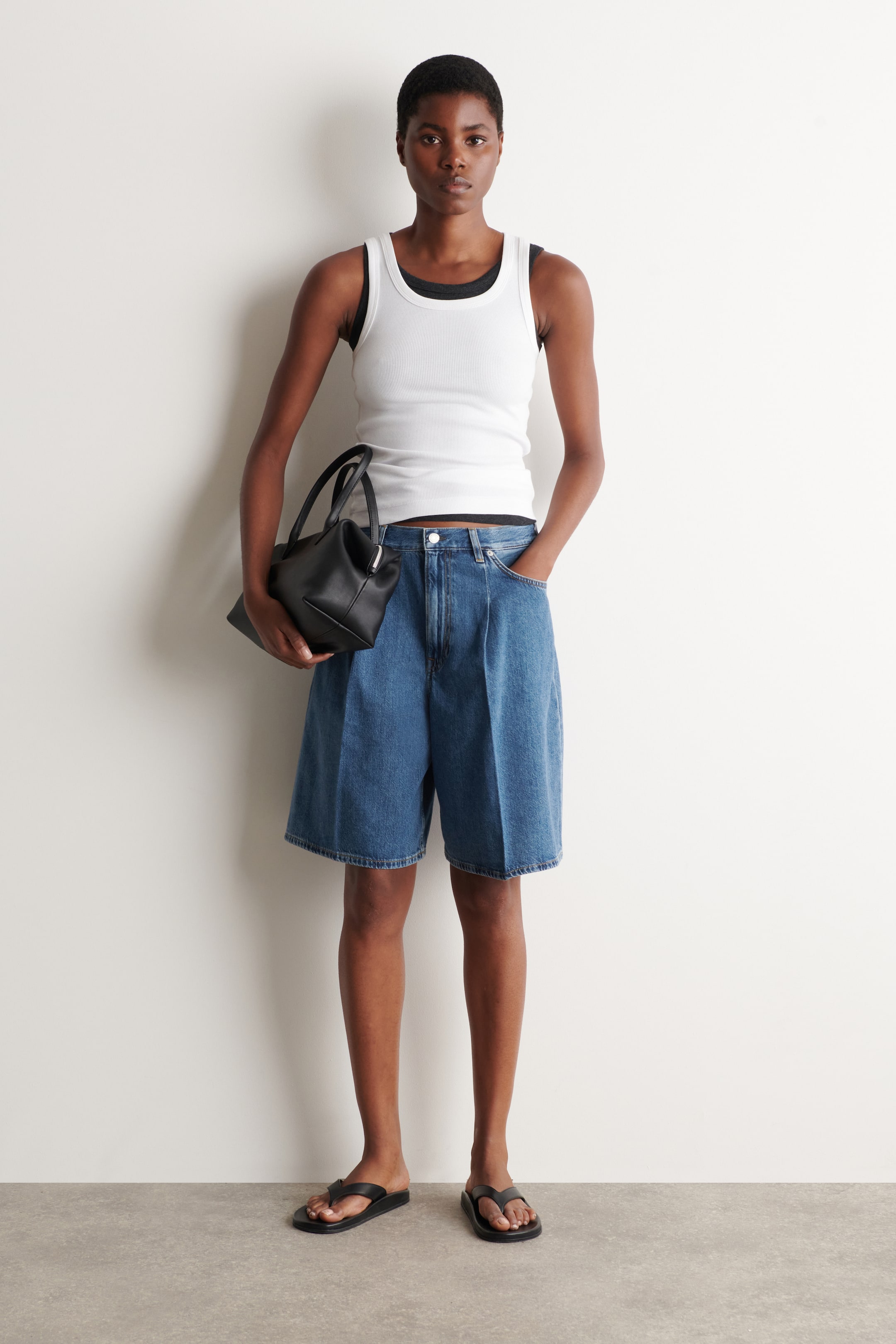 Grösseres Bild ansehen: Blaue Denim-Shorts mit einer entspannten, weiten Passform und Falten werden vom Model getragen und reichen bis zum Knie. Sie verfügen über einen sichtbaren Knopfverschluss. Das Model steht mit einer Hand in der Hosentasche, kombiniert mit einem weißen gerippten Tanktop und einer schwarzen Handtasche.