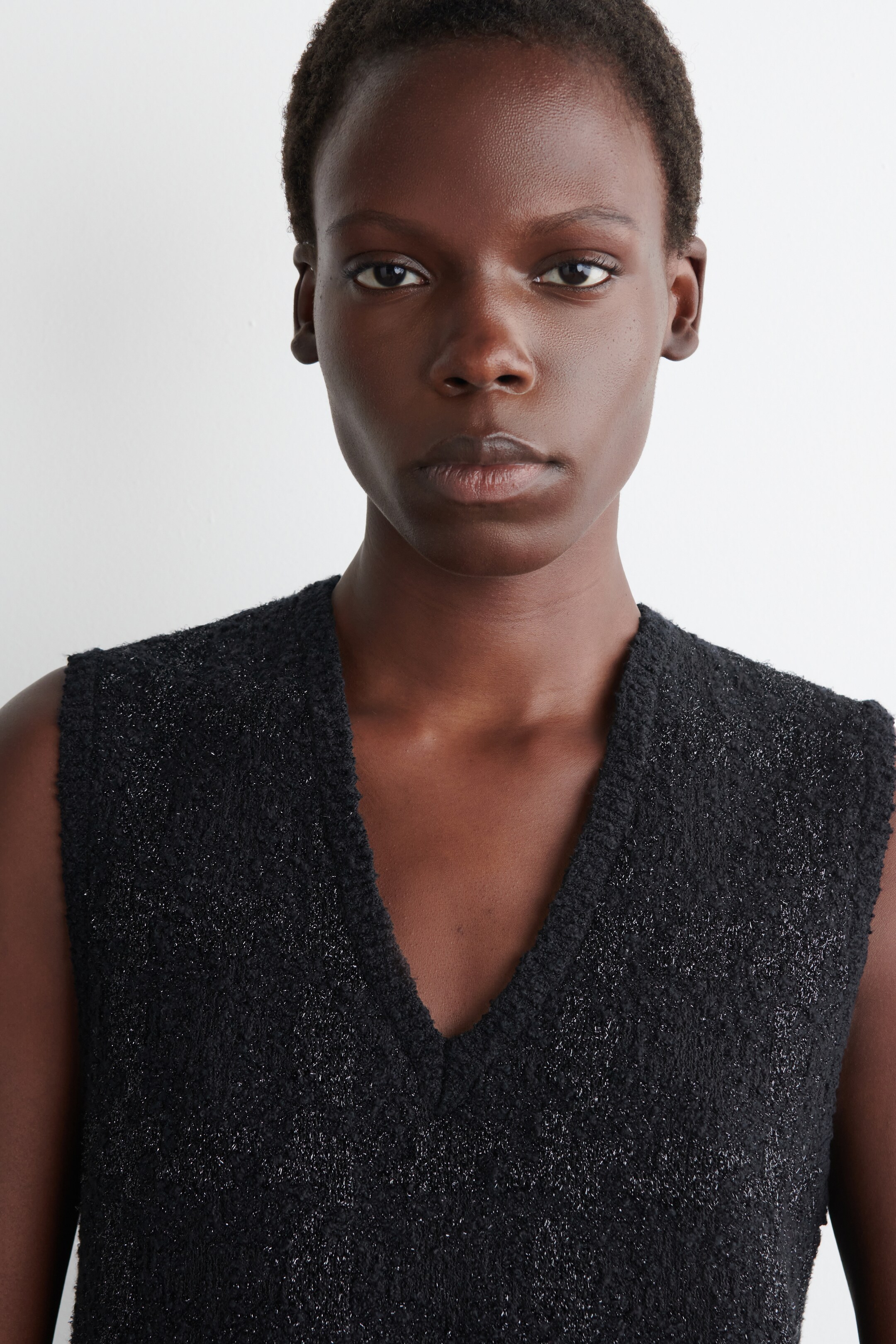 Agrandir l'image: Un haut sans manches en tricot effet bouclé noir est porté, montré de face. Il présente un décolleté en V, et des fils métalliques scintillants sont tissés dans son tissu texturé.