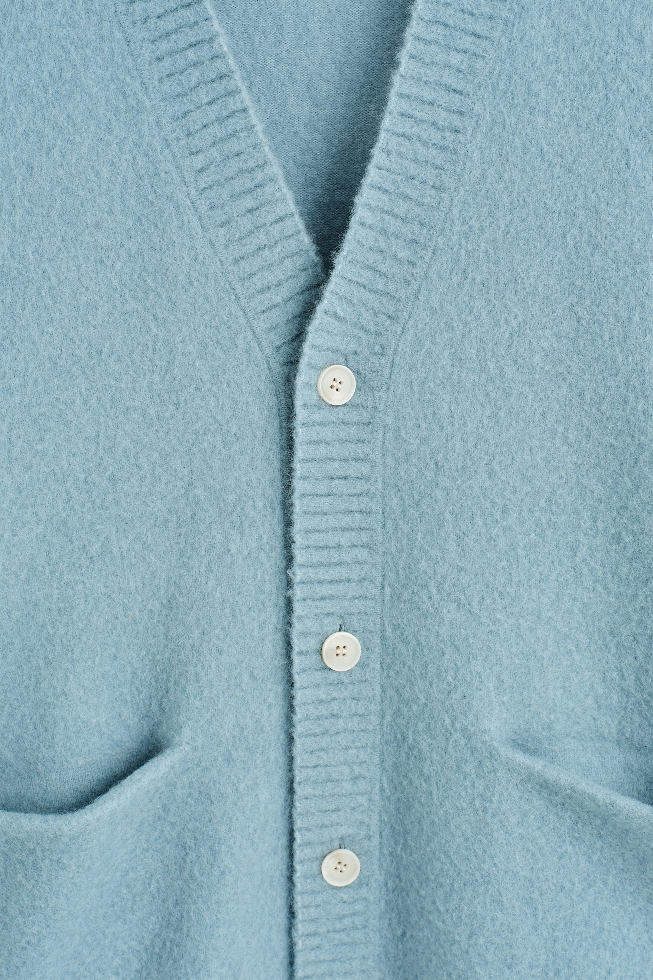 Ver imagen más grande: Un cárdigan azul claro muestra su parte frontal, con una textura cepillada, una solapa de cuello en V acanalada y tres botones de color claro que cierran el centro, con un bolsillo de parche parcialmente visible en la parte inferior delantera.