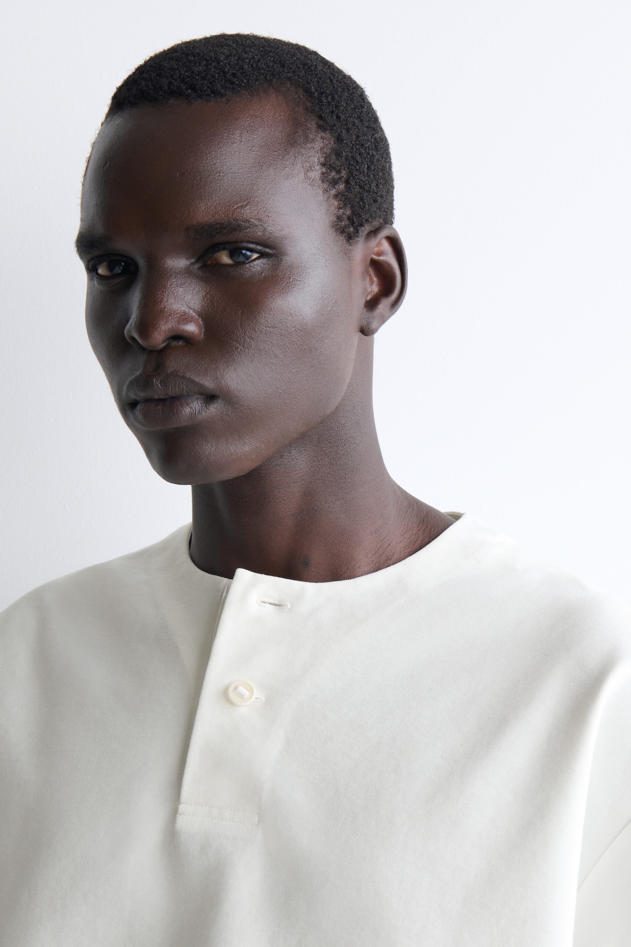 Un t-shirt Henley oversize en jersey de coton est présenté en gros plan, mettant en valeur sa couleur blanc cassé et la texture lisse de son tissu. Le haut présente un col rond sans col avec une patte de boutonnage partielle, montrant un bouton fermé et une boutonnière ouverte.