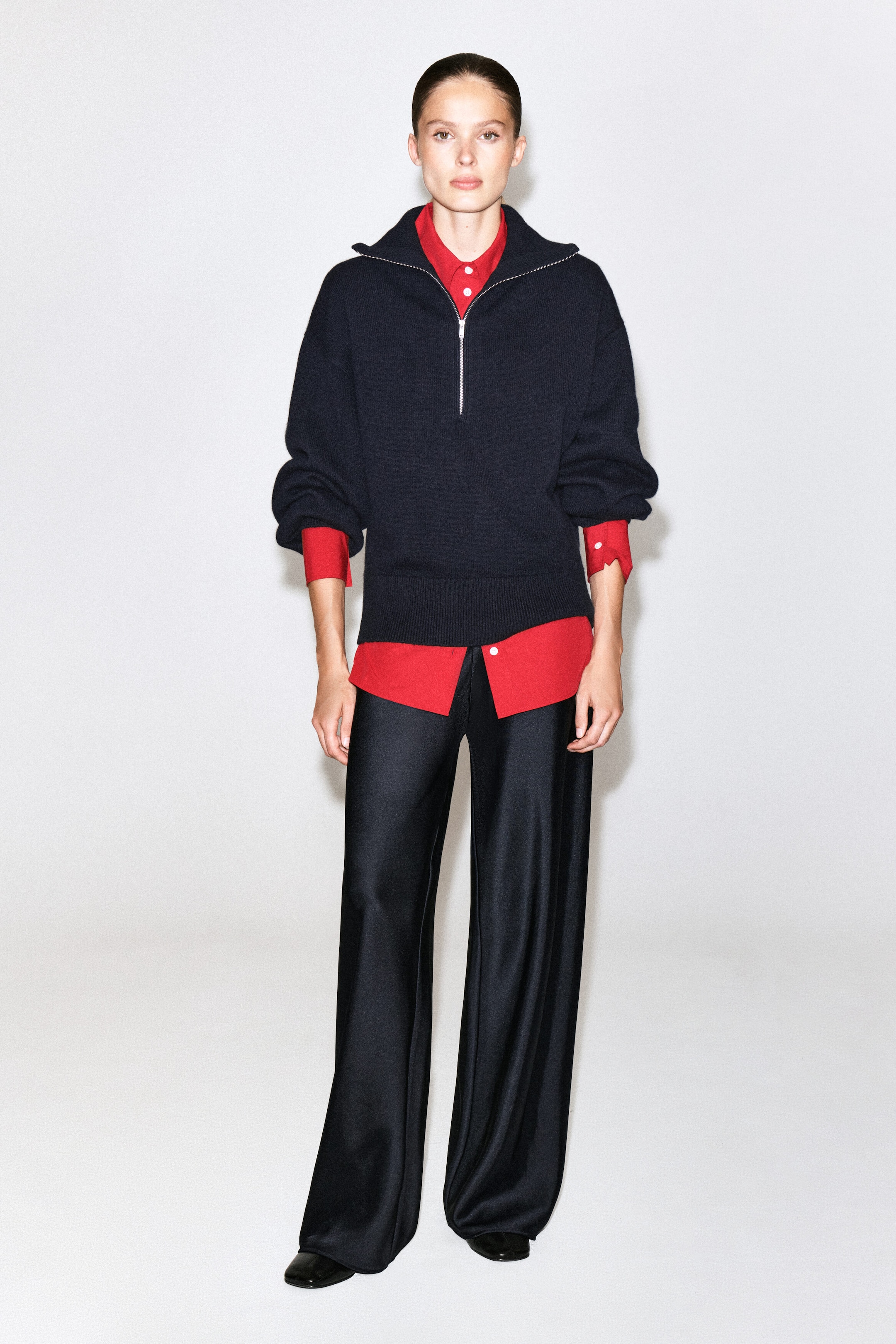 A dark navy half-zip knit jumper is being worn by the model over a bright red collared shirt, paired with wide-leg black satin trousers. The red shirt's collar, cuffs, and hem are visible beneath the jumper, which features a partially open silver zip and ribbed trim.