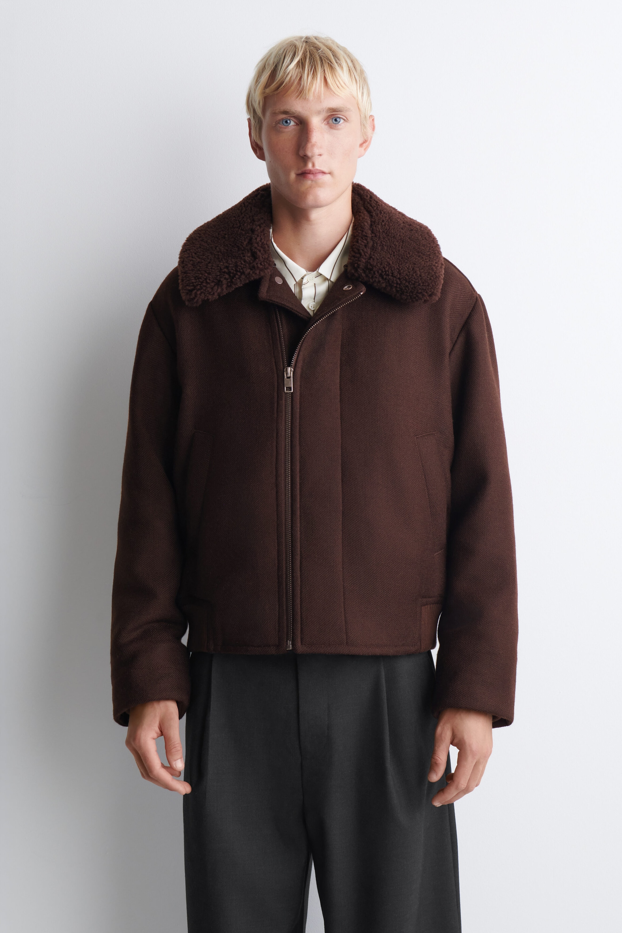This dark brown structured jacket features a plush, textured shearling collar and a silver-tone front zipper. The jacket has a relaxed fit with long sleeves and side welt pockets, layered over a light-colored collared shirt and dark gray trousers. This dark brown structured jacket features a plush, textured shearling collar and a silver-tone front zipper. The jacket has a relaxed fit with long sleeves and side welt pockets, layered over a light-colored collared shirt and dark gray trousers.