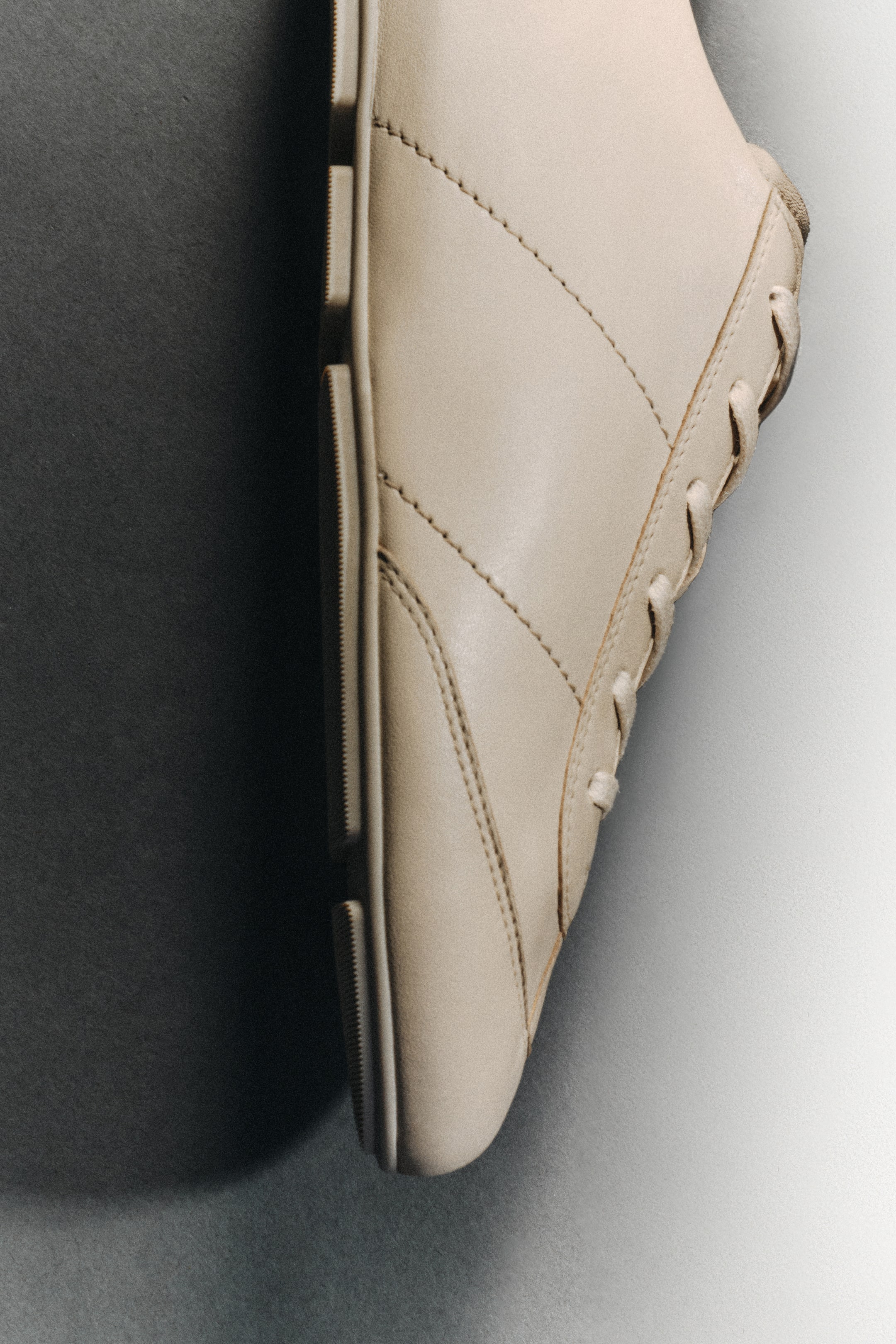 Größeres Bild ansehen: Ein hellbeiger Ledersneaker wird aus einem seitlichen Winkel betrachtet, der sein schlankes Design mit tonalen diagonalen Ziernähten, einem Schnürverschluss mit passenden Schnürsenkeln und einer sichtbaren gerippten Gummisohle zeigt.