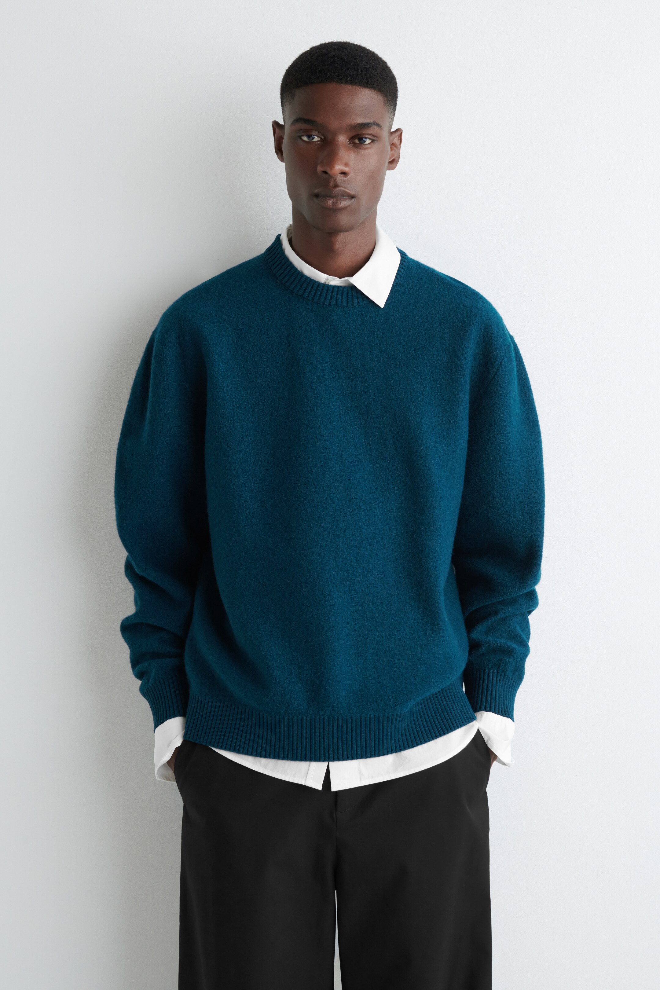 View larger image: A teal crewneck sweater with a soft, fuzzy texture is being shown from the front, presented in a relaxed fit. It is featuring ribbed details on the collar, cuffs, and hem, and is being layered over a white collared shirt.