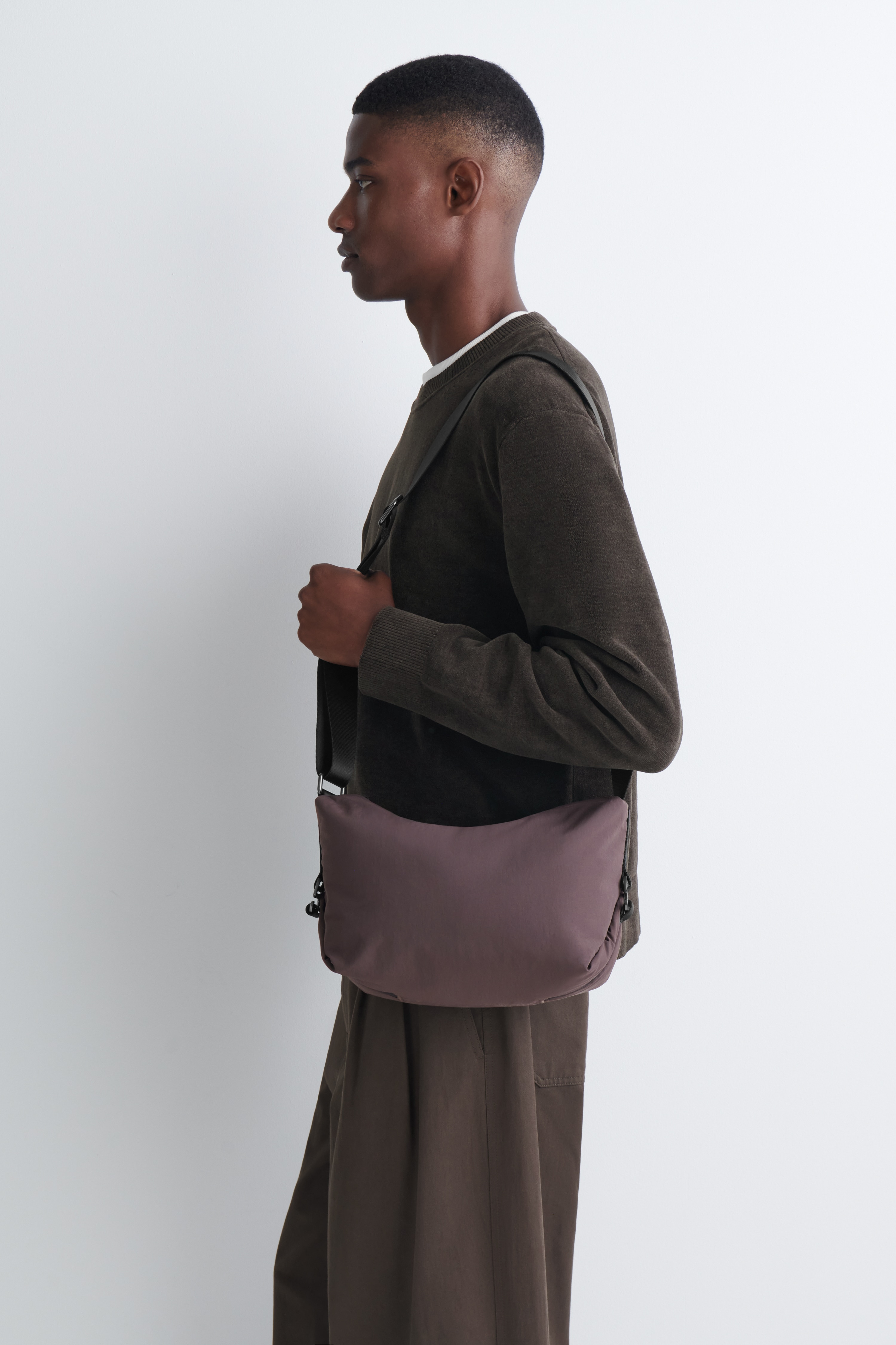 A compact, rectangular crossbody bag in dusty plum technical nylon is shown from the front, hanging from its long, adjustable black webbing shoulder strap. The minimalist bag features webbing straps that fasten with black buckles on each side.