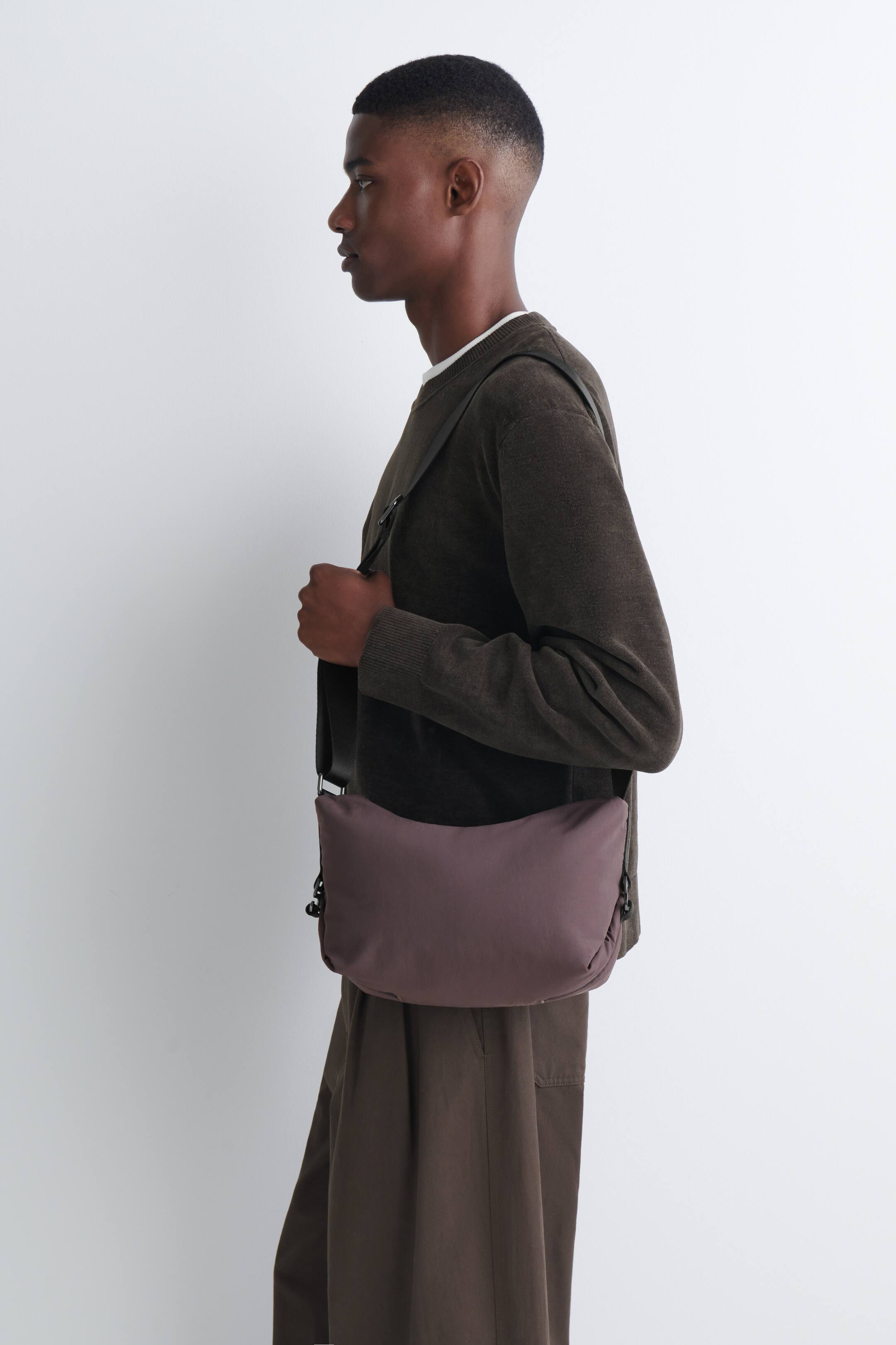 View larger image: A mauve-brown nylon crossbody bag is worn over the shoulder by the model, who is holding the dark adjustable strap. The compact, slouchy bag features side buckle details and is styled with a dark green long-sleeved jumper and brown trousers.