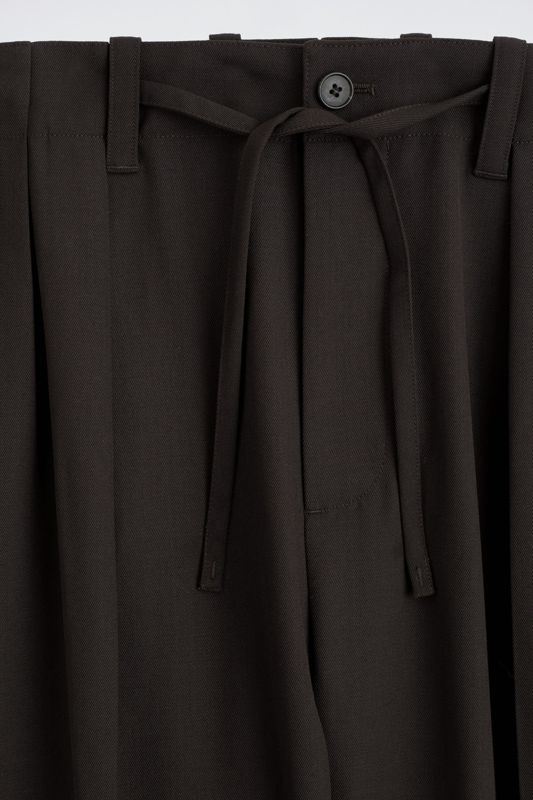 Dark brown trousers feature a front waistband with a single button closure, belt loops, and a matching fabric tie belt knotted at the centre. The trousers showcase a textured twill weave with soft pleats draping downward. Dark brown trousers feature a front waistband with a single button closure, belt loops, and a matching fabric tie belt knotted at the centre. The trousers showcase a textured twill weave with soft pleats draping downward.