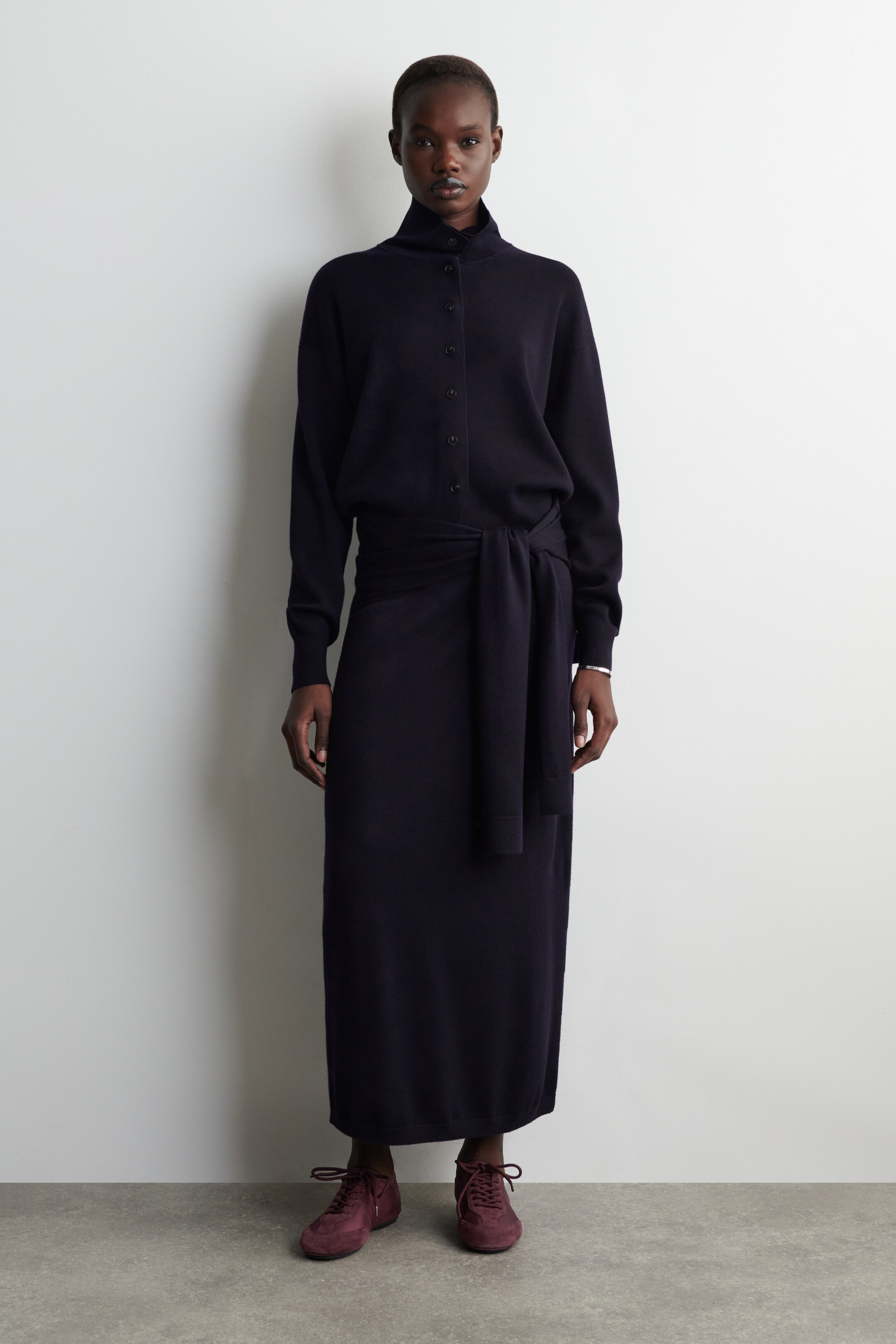 Agrandir l'image: Une robe à manches longues bleu marine foncé est présentée, face avant. Elle est dotée d'un col montant et d'une patte de boutonnage sur le corsage. Une ceinture en tissu assorti est nouée autour de la taille, et la robe se prolonge en une jupe droite, longueur cheville.