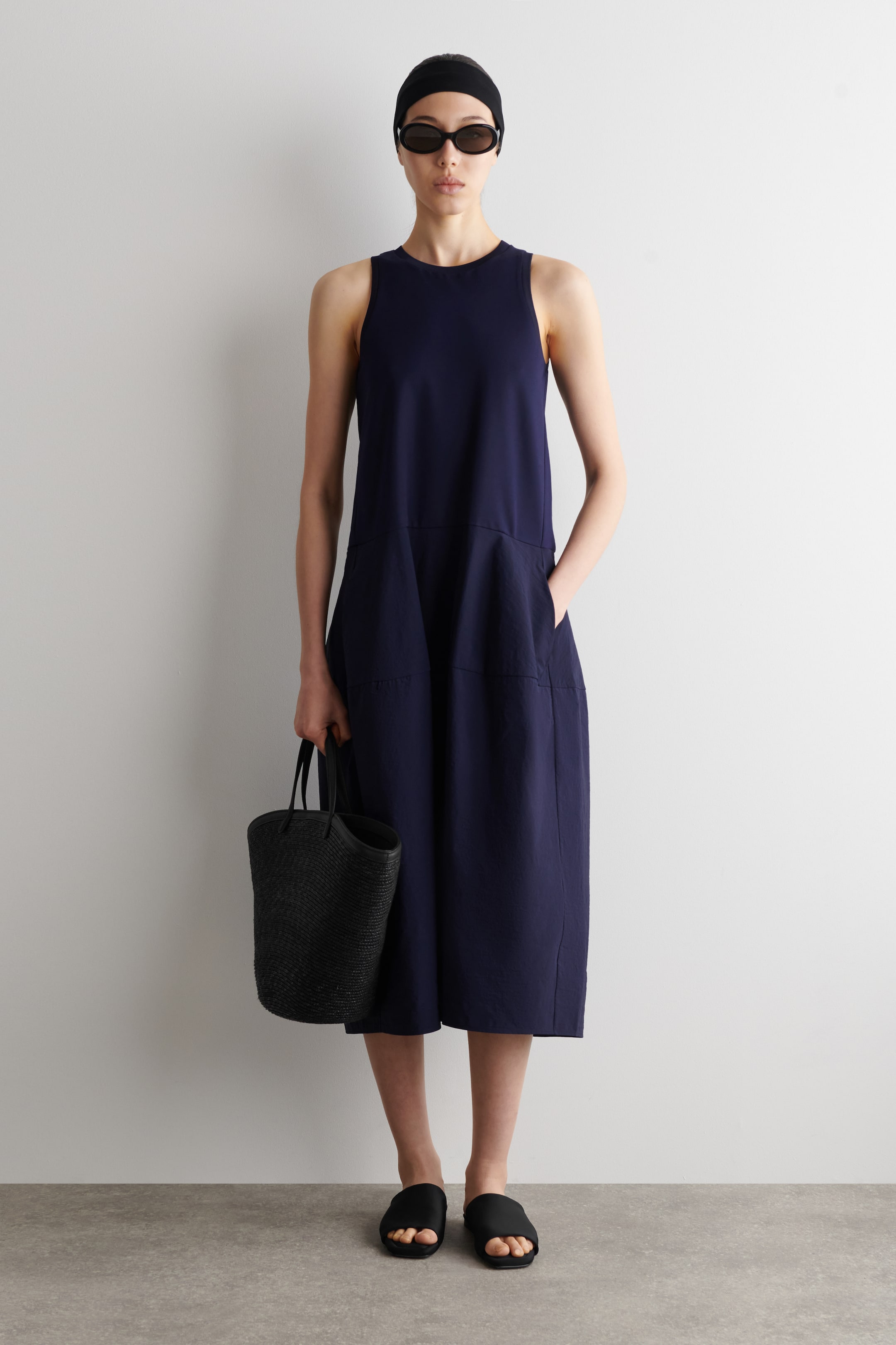 Visualizza l'immagine ingrandita: Un abito blu navy presenta un design senza maniche con scollo rotondo, offrendo una silhouette rilassata di lunghezza midi che si allarga dalla vita, con due tasche applicate sul davanti.