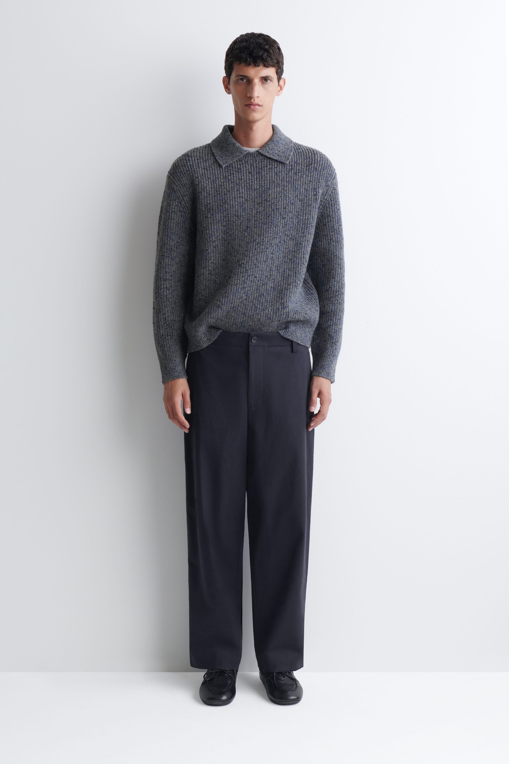 A grey, ribbed-knit sweater featuring a polo-style collar and long sleeves is being worn by the model. The relaxed-fit sweater is styled tucked into dark, wide-leg trousers and paired with black shoes. A grey, ribbed-knit sweater featuring a polo-style collar and long sleeves is being worn by the model. The relaxed-fit sweater is styled tucked into dark, wide-leg trousers and paired with black shoes.