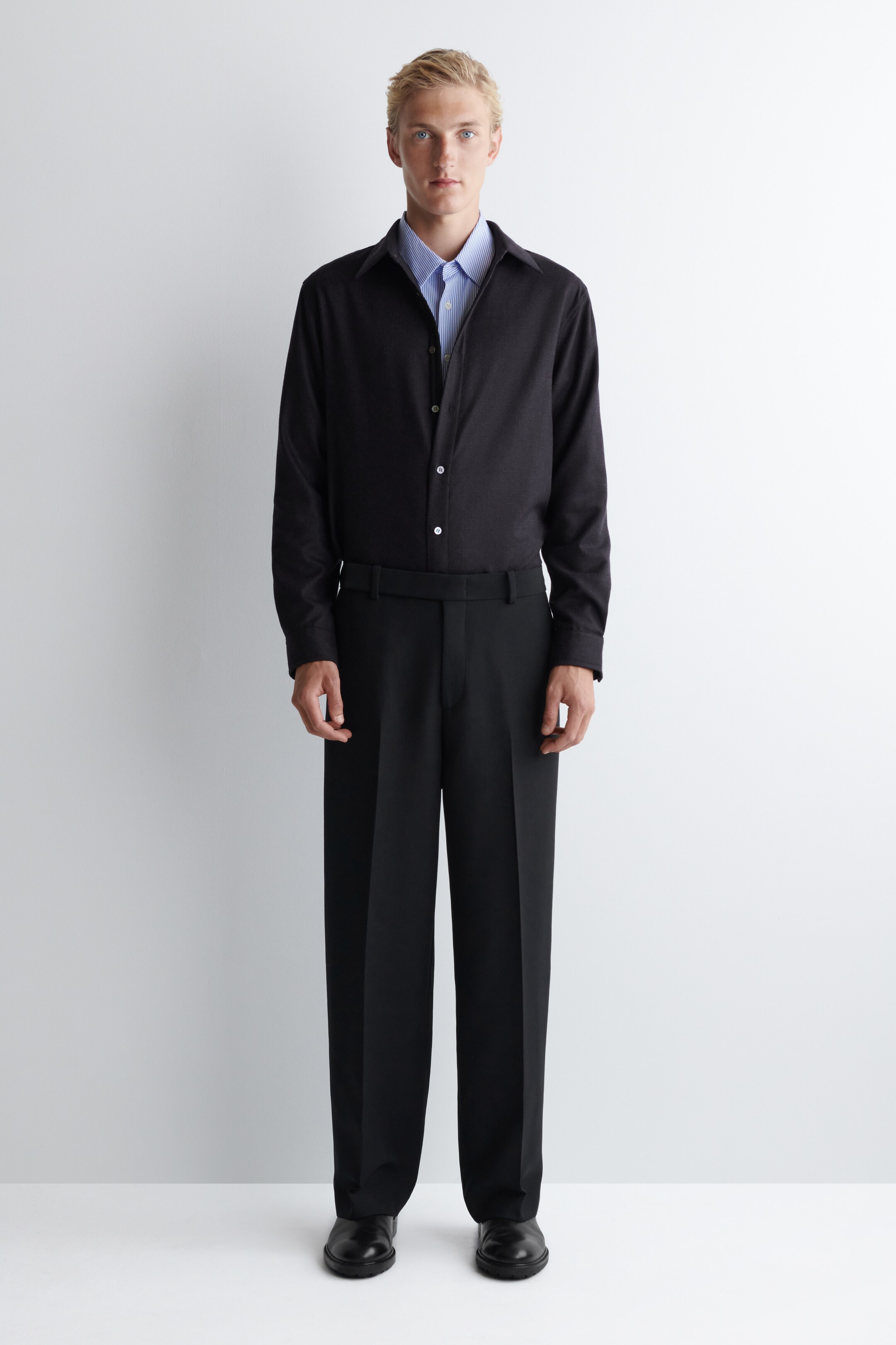 View larger image: A dark long-sleeved button-up shirt, displaying white buttons down its front, is being worn with its collar open over a light blue striped shirt. Wide-leg black trousers with a flat front and belt loops are being worn, paired with black rounded-toe shoes.