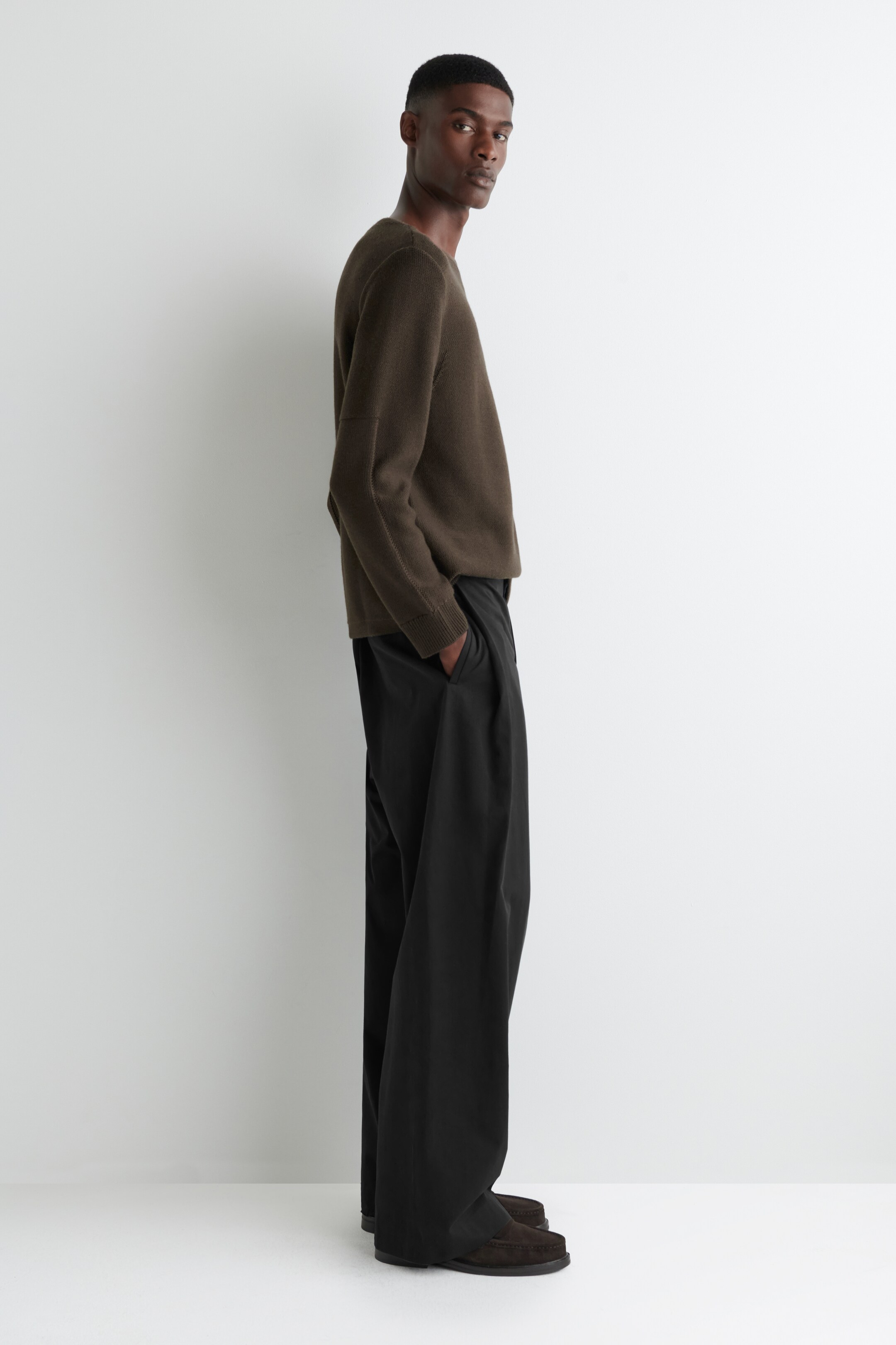 Agrandir l'image: Un pull à col rond vert olive avec des coutures apparentes sur les manches et des poignets côtelés est porté. Il est assorti à un pantalon noir à jambes larges et plis, qui s'accumule aux chevilles, et des mocassins marron foncé, le tout présenté en vue de profil latéral.