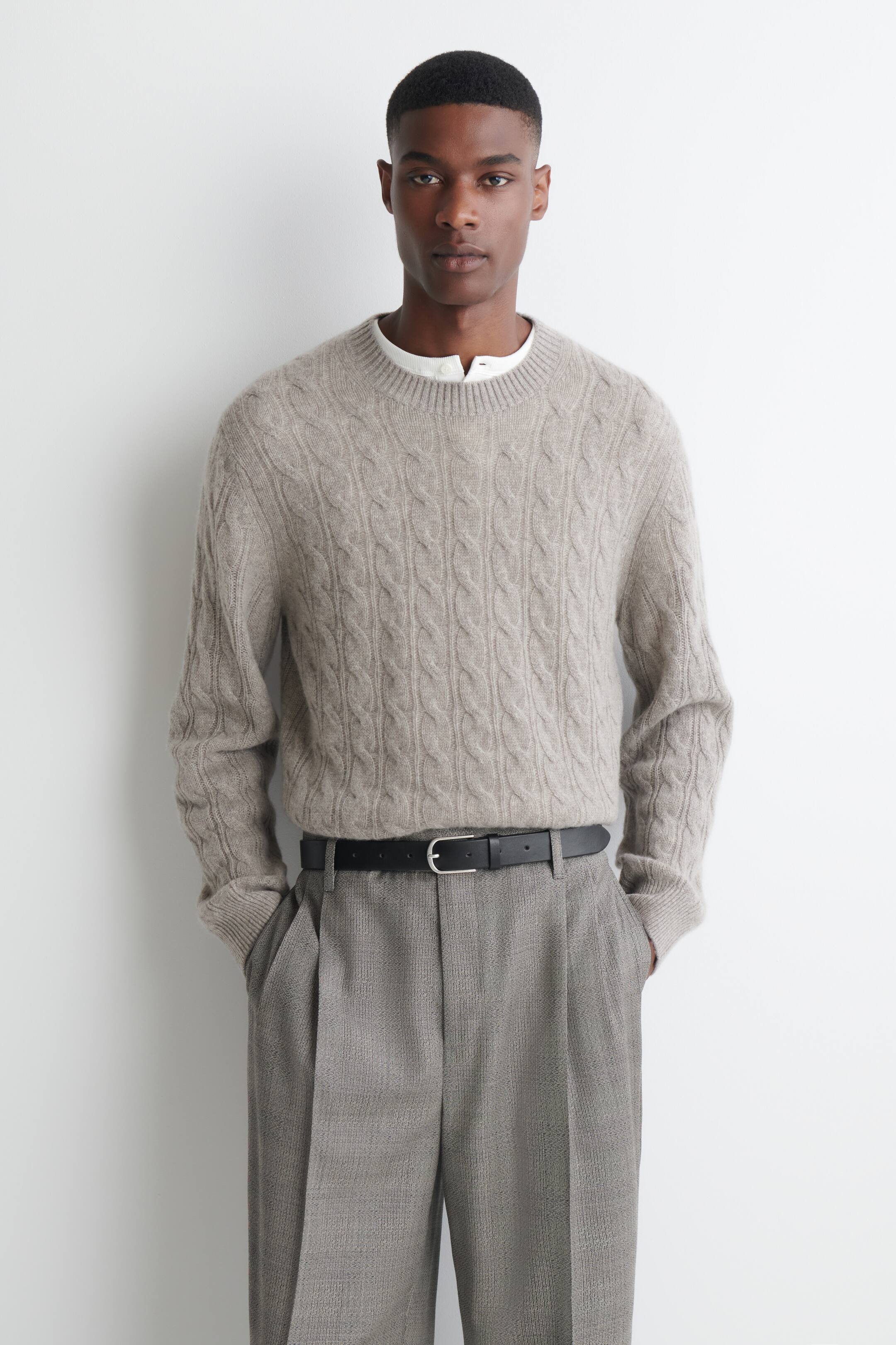 Visualizza l'immagine ingrandita: Un maglione girocollo in cashmere lavorato a trecce, di colore beige chiaro, è indossato dalla modella sopra una camicia bianca con colletto e bottoni. Il maglione, caratterizzato da un motivo a trecce su tutta la superficie e finiture a coste, è infilato in pantaloni grigi con pieghe, tenuti in vita da una cintura nera.
