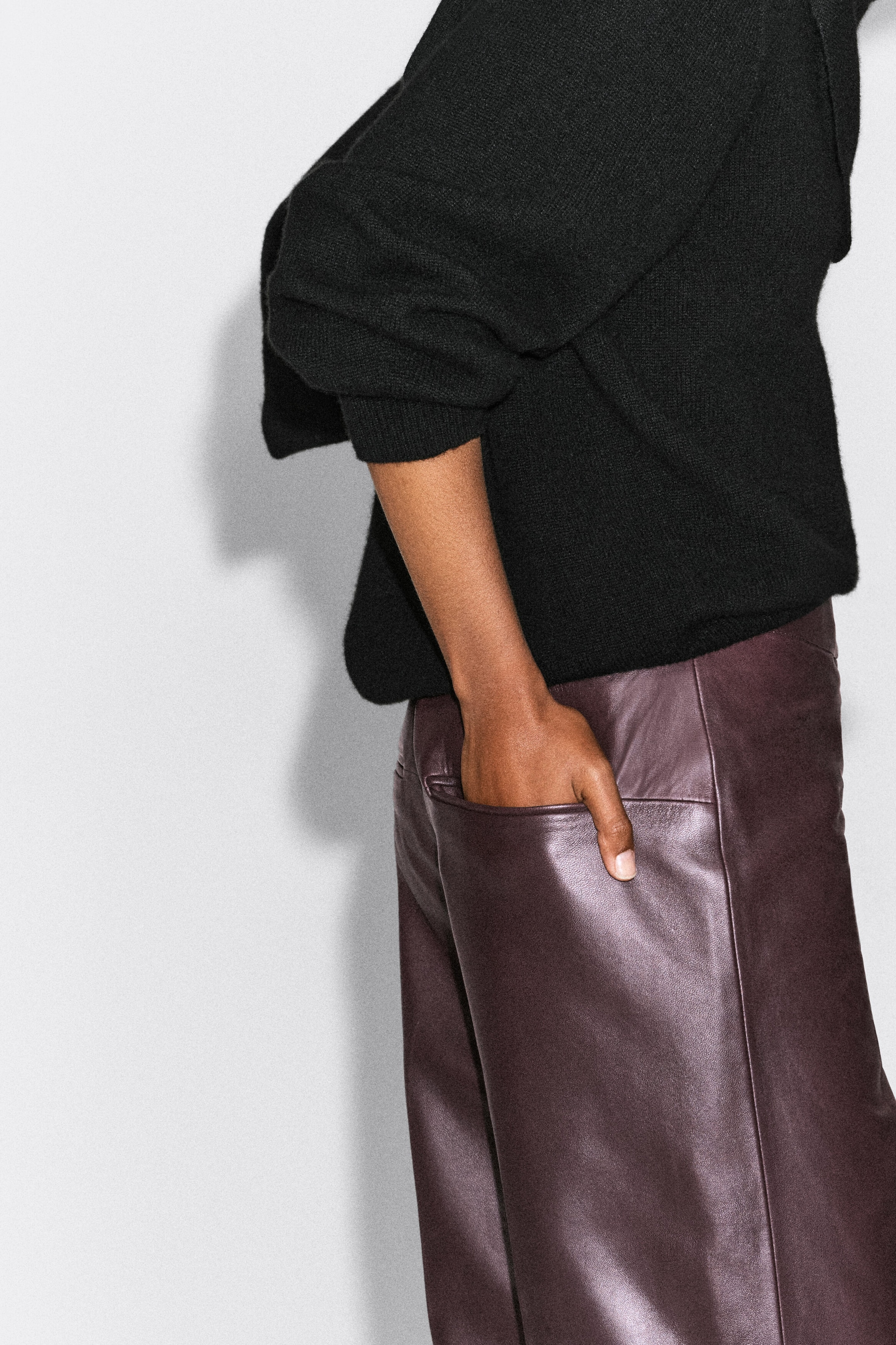 Leather straight-leg pants in a deep burgundy color are being worn by the model, who is resting a hand in the back pocket. The pants feature a smooth leather texture and are paired with a black knit sweater. Leather straight-leg pants in a deep burgundy color are being worn by the model, who is resting a hand in the back pocket. The pants feature a smooth leather texture and are paired with a black knit sweater.