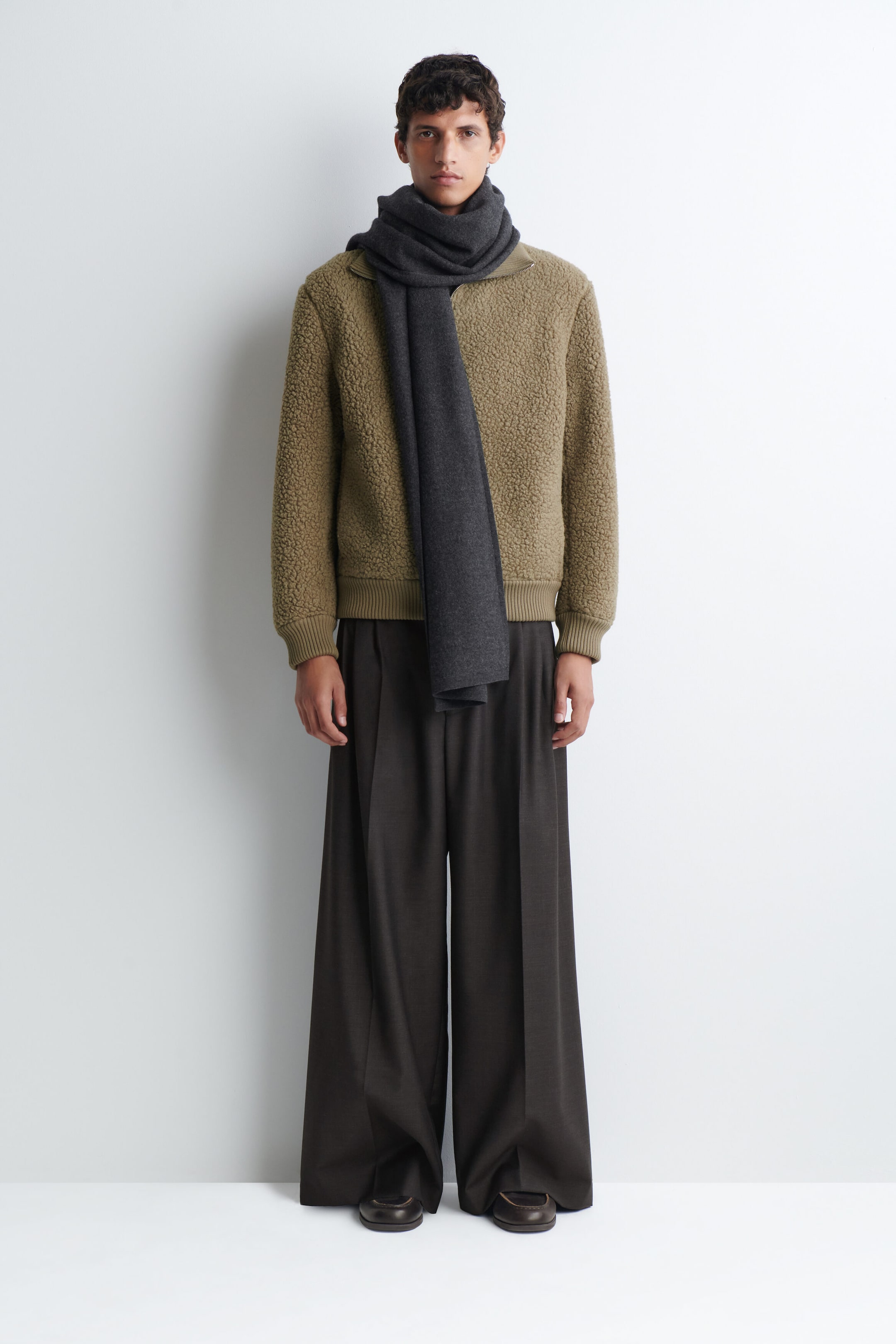 The model is wearing an olive green fleece jacket with a high collar, ribbed cuffs, and a ribbed hem. The jacket is styled with a long, dark grey scarf wrapped around the neck and dark brown, wide-leg pleated trousers. The model is wearing an olive green fleece jacket with a high collar, ribbed cuffs, and a ribbed hem. The jacket is styled with a long, dark grey scarf wrapped around the neck and dark brown, wide-leg pleated trousers.