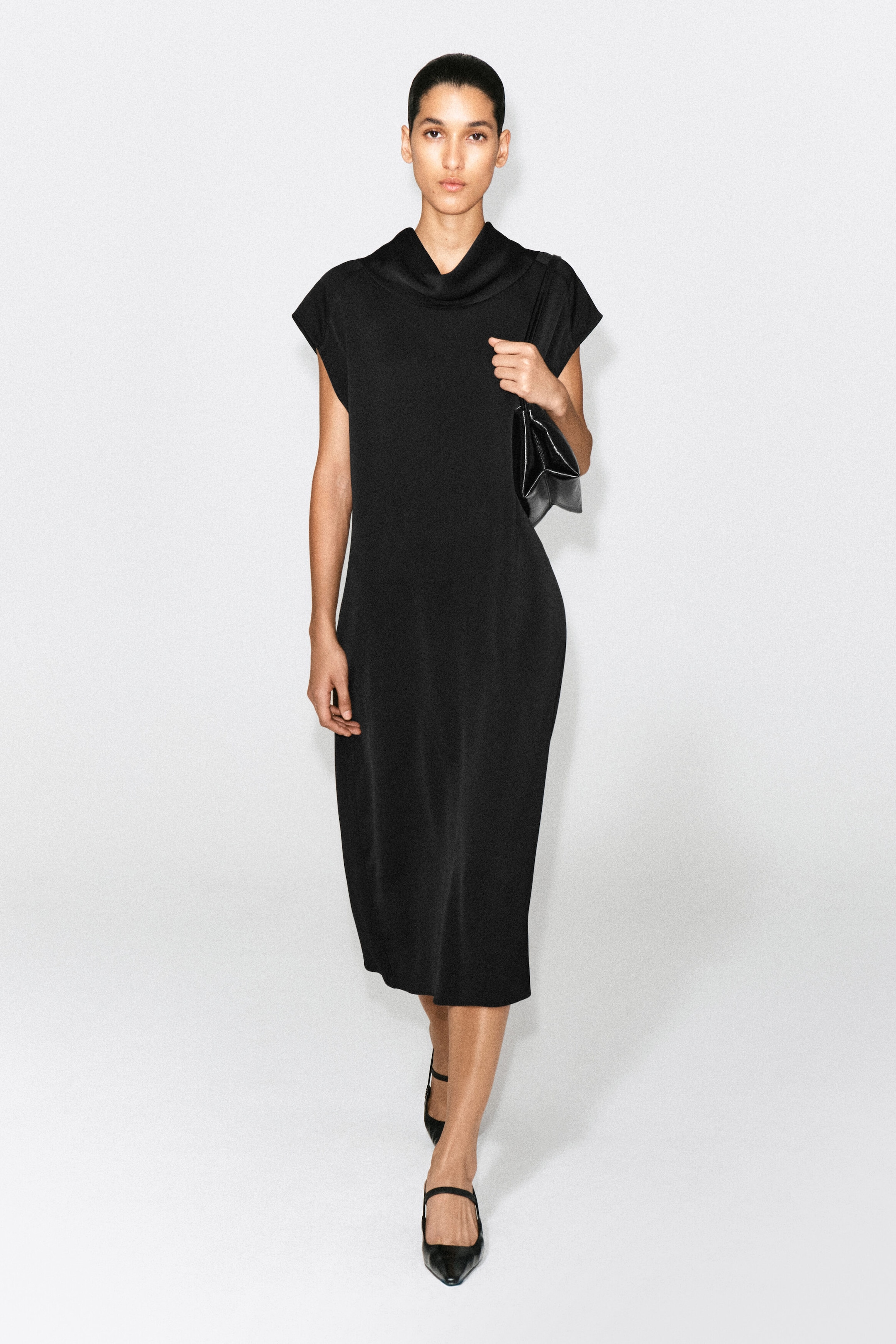 A black cowl-neck crepe midi dress is shown from the front, featuring a relaxed silhouette with extended shoulders that create cap sleeves. The dress drapes softly at the neckline and falls to a midi length.
