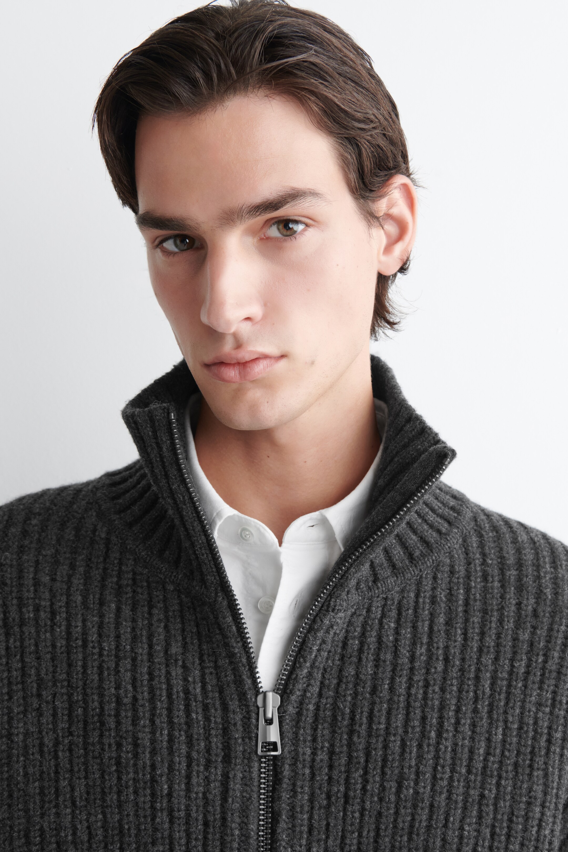 Agrandir l'image: Un cardigan zippé côtelé en tricot gris foncé est présenté de face, avec un col montant et une fermeture éclair argentée sur toute la longueur, porté par-dessus une chemise blanche à col.