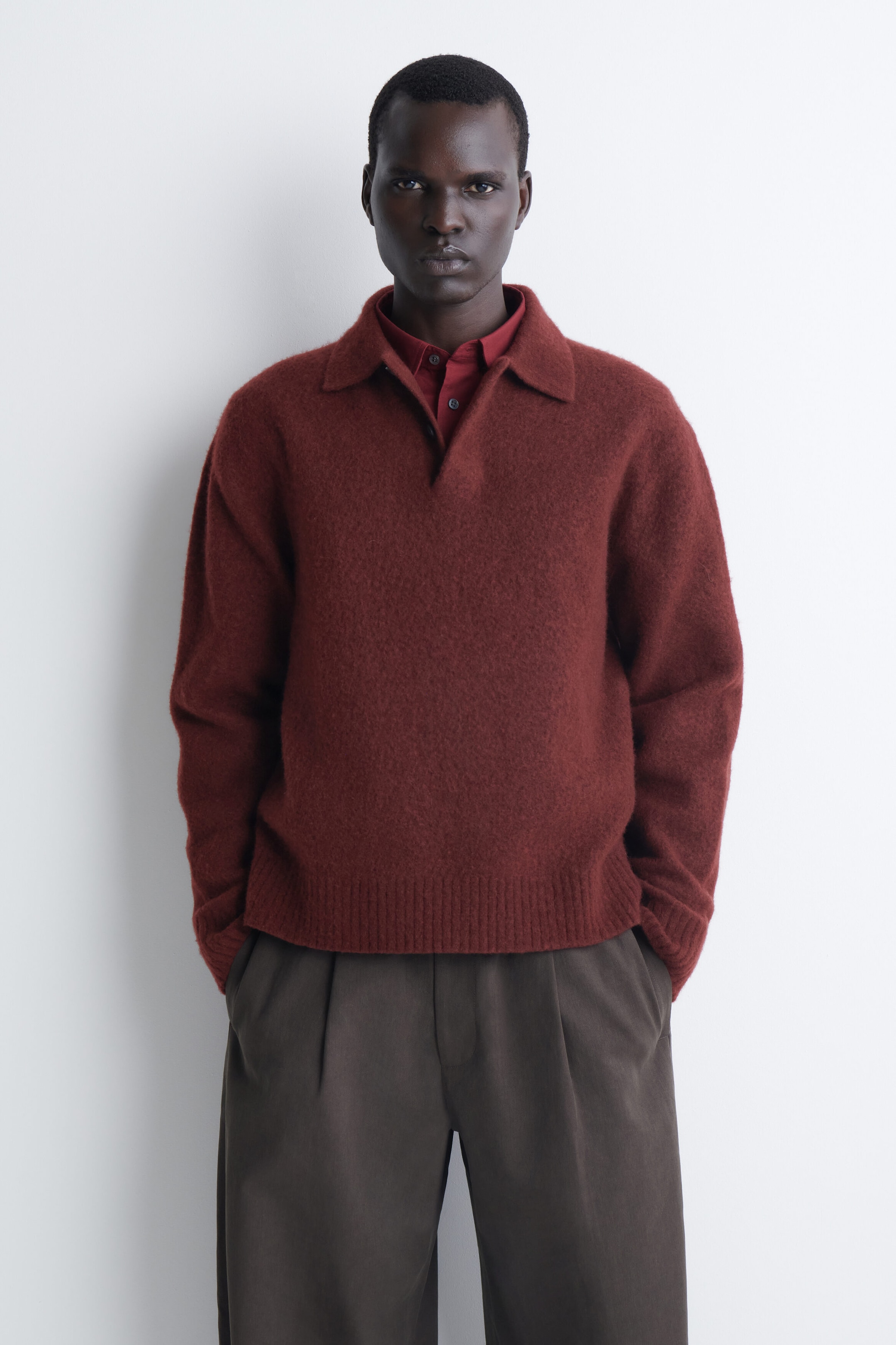 The brushed wool polo shirt in a reddish-brown shade is shown from the front, featuring a soft, textured finish, a classic polo collar, and ribbed detailing at the hem and cuffs. The shirt has a relaxed fit and is layered over a dark red button-up collared shirt. The brushed wool polo shirt in a reddish-brown shade is shown from the front, featuring a soft, textured finish, a classic polo collar, and ribbed detailing at the hem and cuffs. The shirt has a relaxed fit and is layered over a dark red button-up collared shirt.