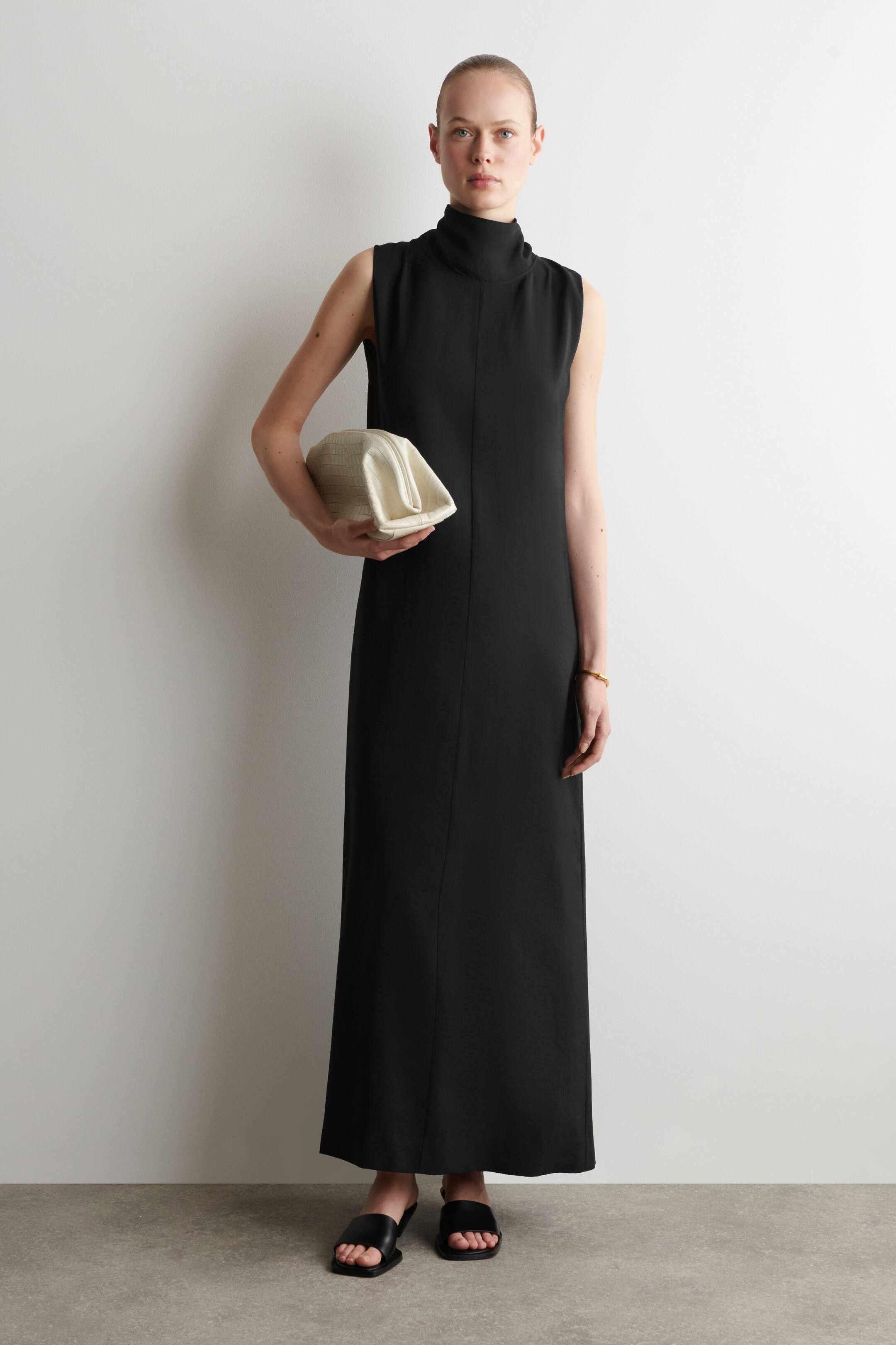 Agrandir l'image: Une robe maxi noire présente un col montant et un design sans manches. La robe tombe droit jusqu'aux chevilles, et une couture verticale subtile parcourt le centre avant.