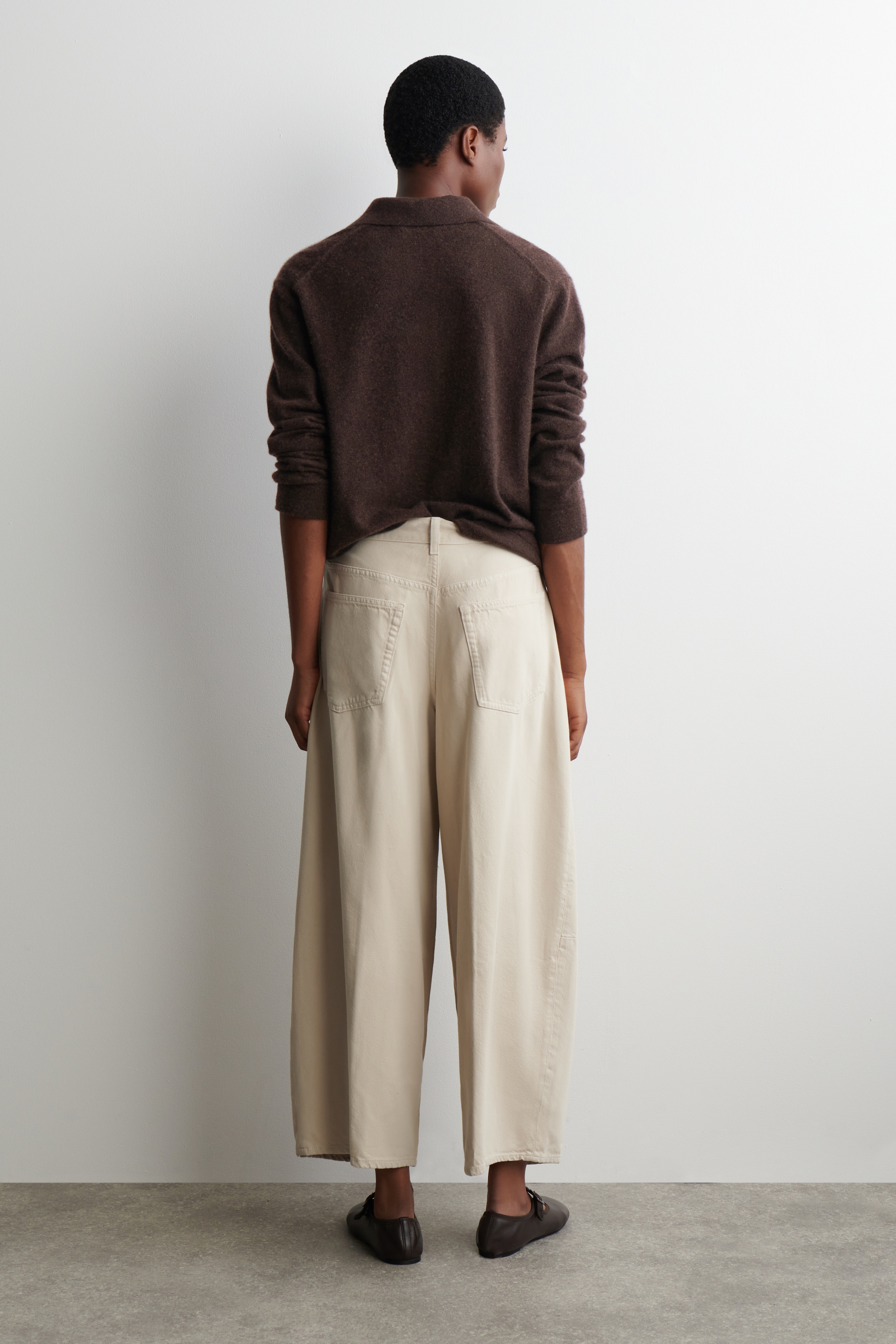 View larger image: A dark brown long-sleeved collared top is being worn, presented from the back. It is paired with light beige wide-leg pants, which are featuring visible back pockets and are styled with dark brown buckled flat shoes.