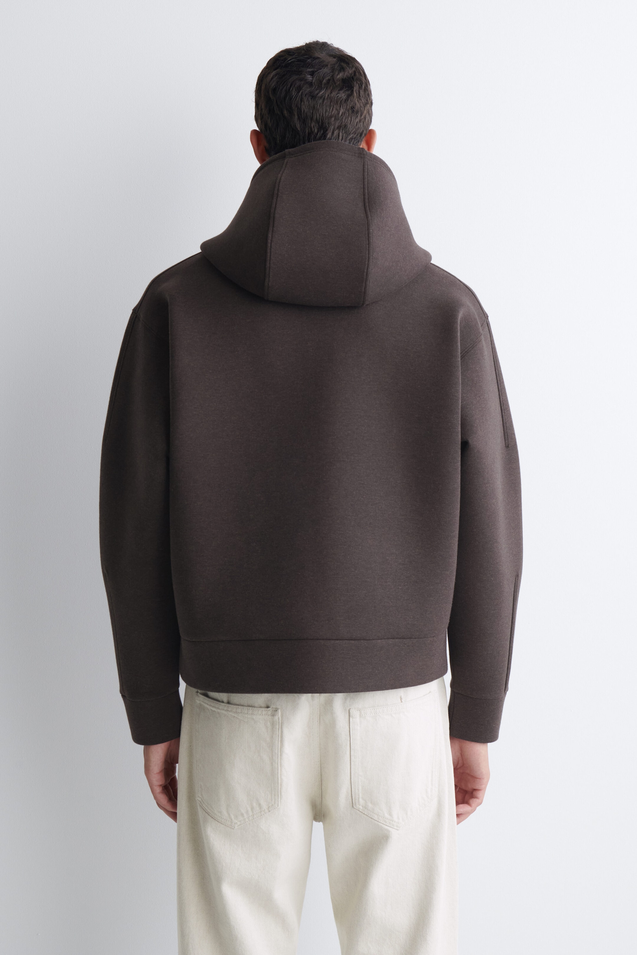 An oversized dark brown scuba jersey hoodie is worn by the model, shown from the back. The hoodie features a structured hood, dropped shoulders, and a ribbed hem, and is styled with light cream-coloured trousers. An oversized dark brown scuba jersey hoodie is worn by the model, shown from the back. The hoodie features a structured hood, dropped shoulders, and a ribbed hem, and is styled with light cream-coloured trousers.