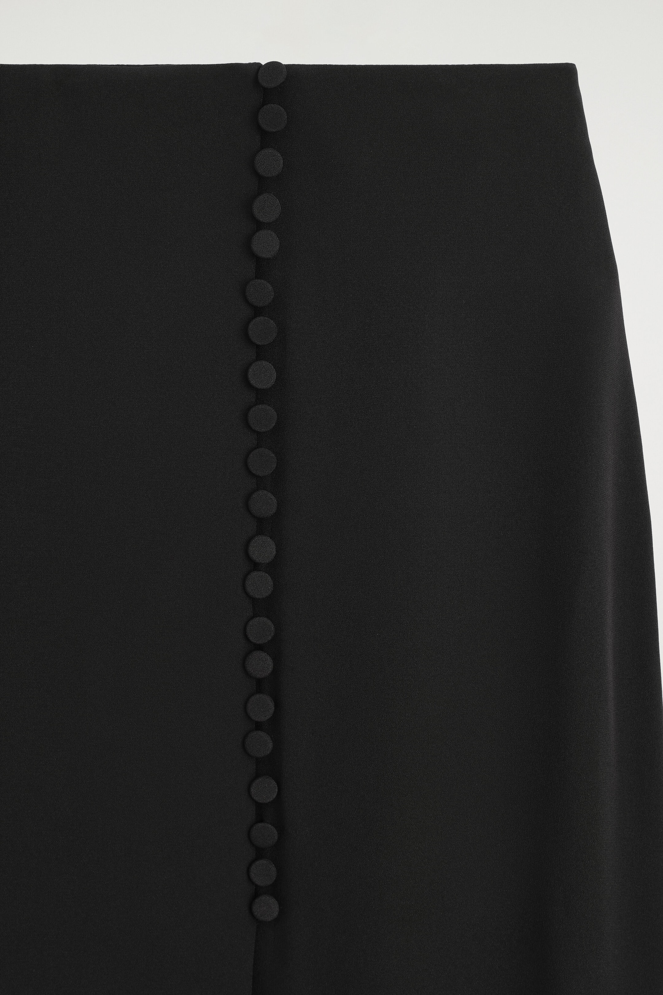 A black skirt is showcasing a vertical row of small, fabric-covered buttons running down its centre front. The smooth, matte fabric is extending downwards from a clean waistline.