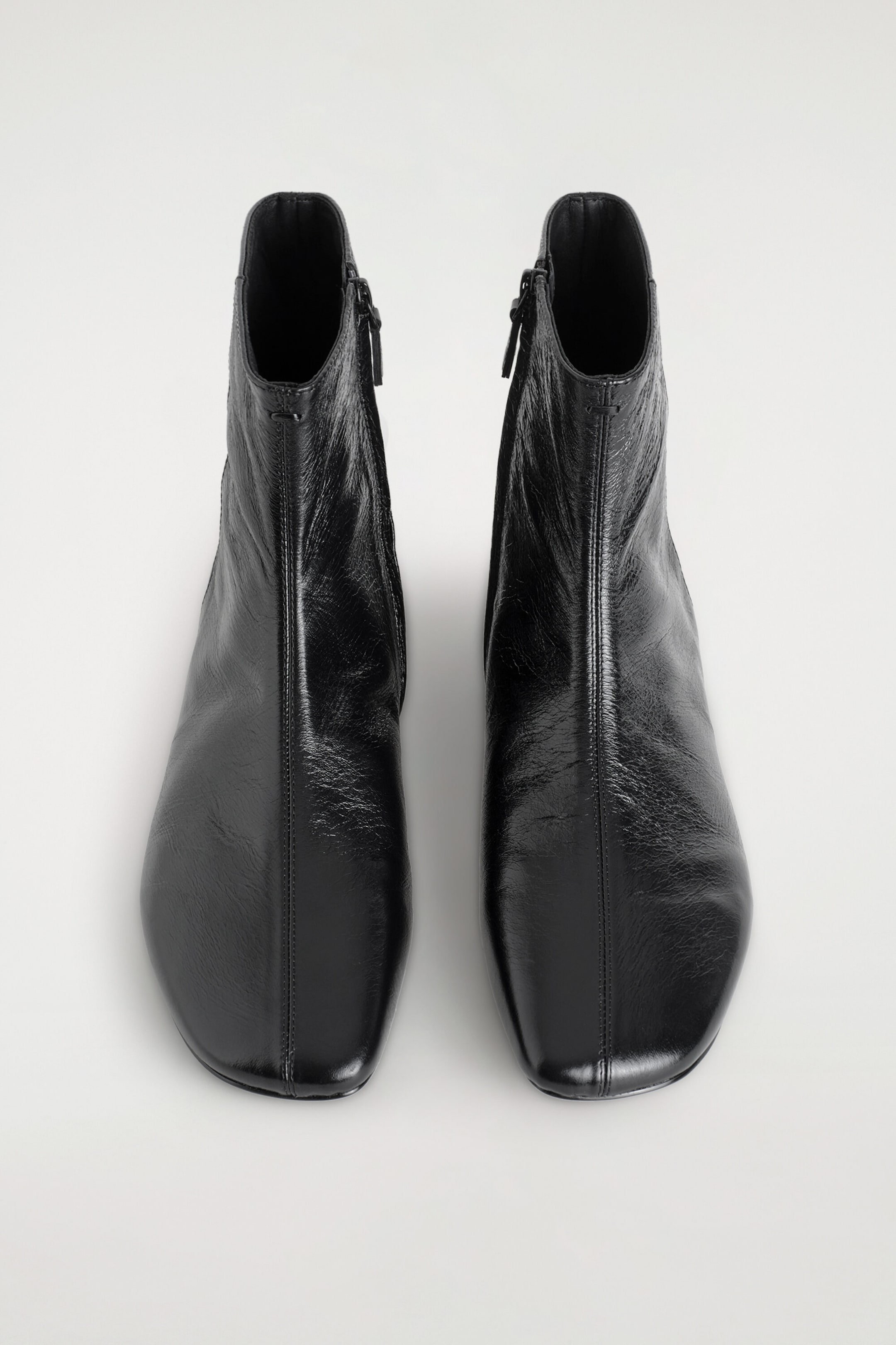 A pair of black leather ankle boots is resting side by side, viewed from above to show their structured square-toe design and glossy, crinkled texture. Each boot features a prominent central vertical seam running down the front and a zip fastening along the inner ankle.