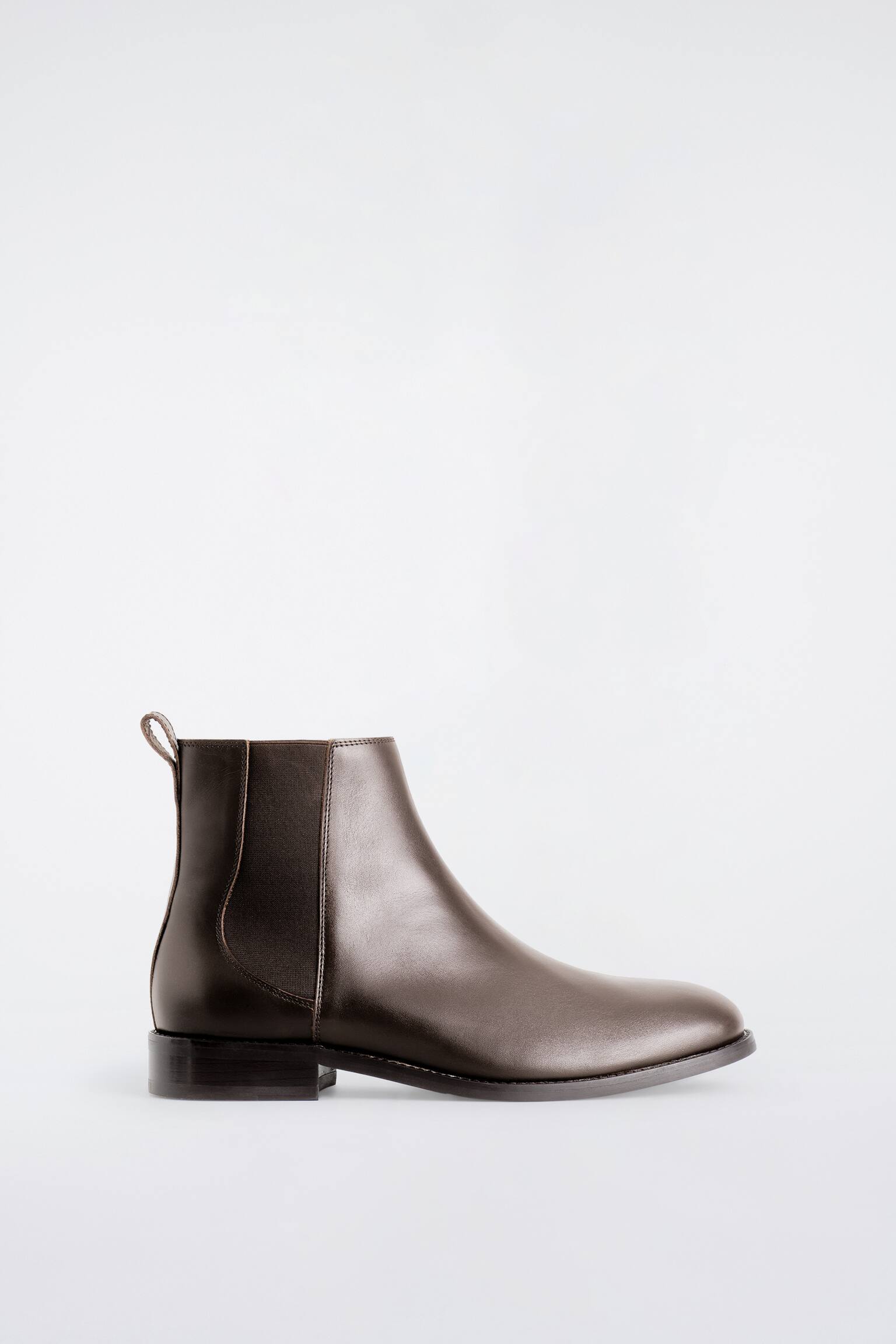 Een donkerbruine leren Chelsea-boot is te zien in zijaanzicht, met een ronde neus, bijpassende elastische zijpanelen en een treklipje aan de achterkant. De boot staat op een lage blokhak en heeft een dunne zool.