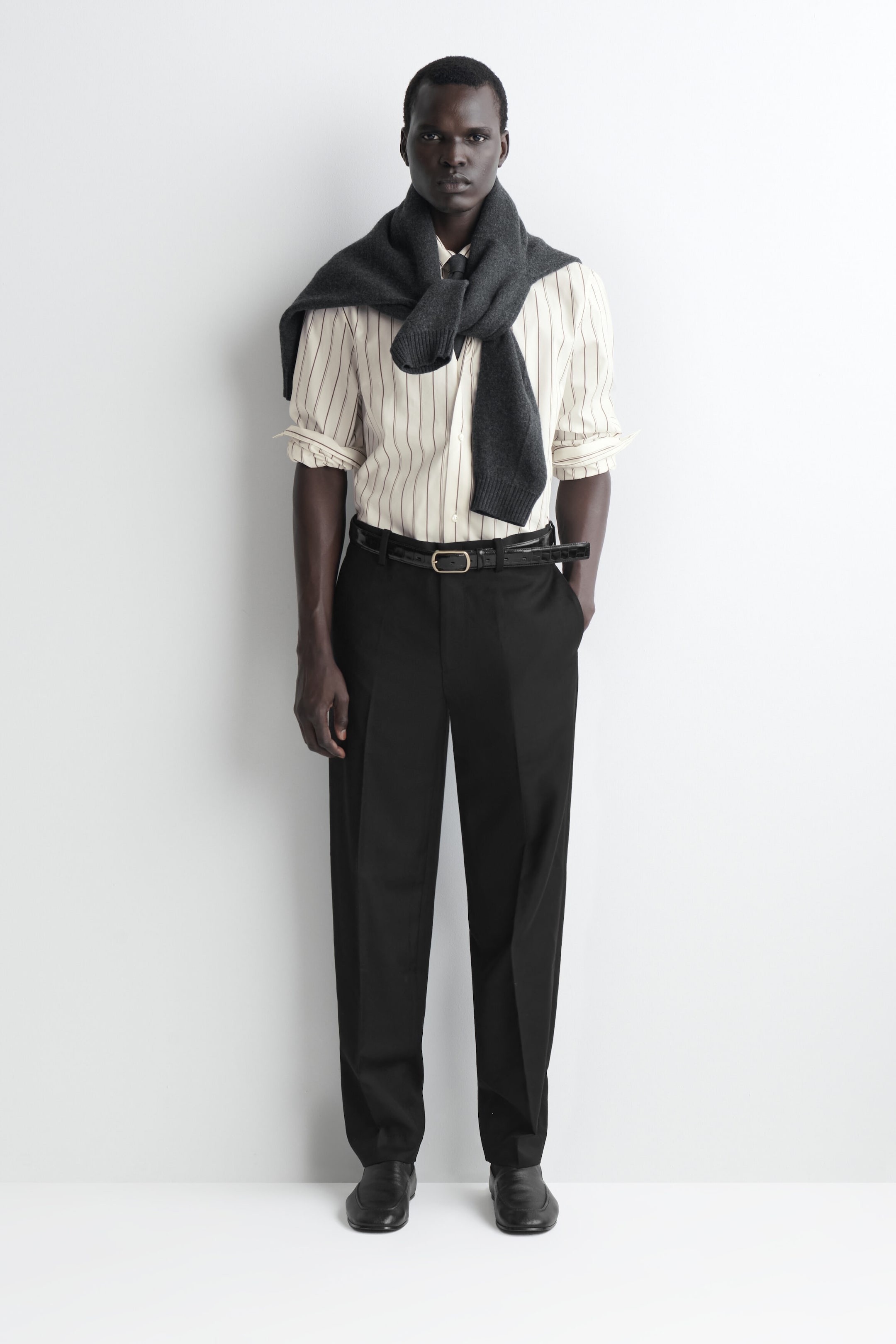 The black wool trousers are shown from the front, featuring a straight-leg silhouette with sharp centre creases down each leg. They are styled with a black leather belt, a tucked-in striped shirt, and black shoes to highlight their regular fit and full-length hem. The black wool trousers are shown from the front, featuring a straight-leg silhouette with sharp centre creases down each leg. They are styled with a black leather belt, a tucked-in striped shirt, and black shoes to highlight their regular fit and full-length hem.