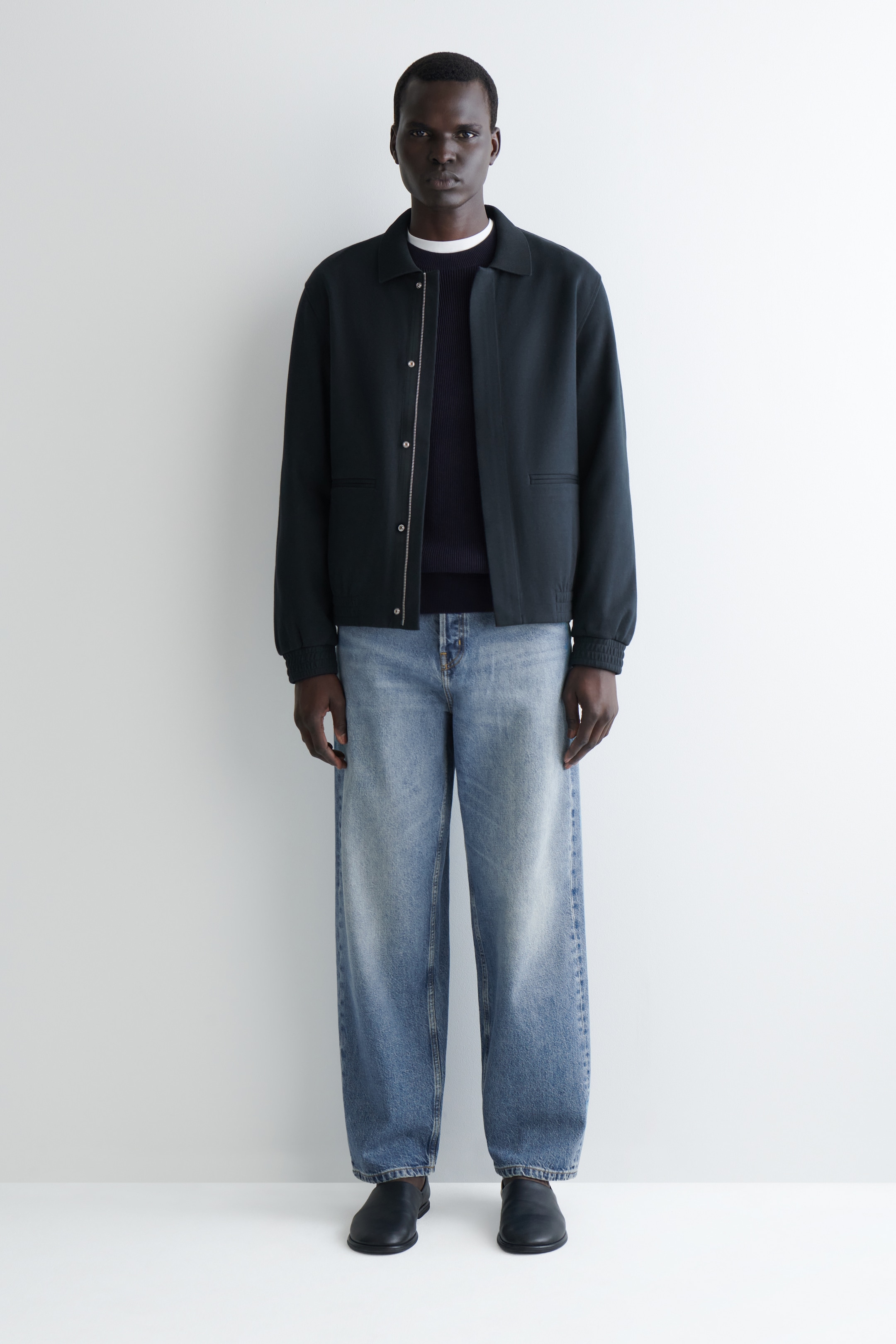 Se større billede: En mørk jakke med krave og elastiske manchetter og kant bæres åbent over en mørk crewneck sweater. Den er parret med lyseblå, løstsiddende jeans og sorte flade slip-on sko.