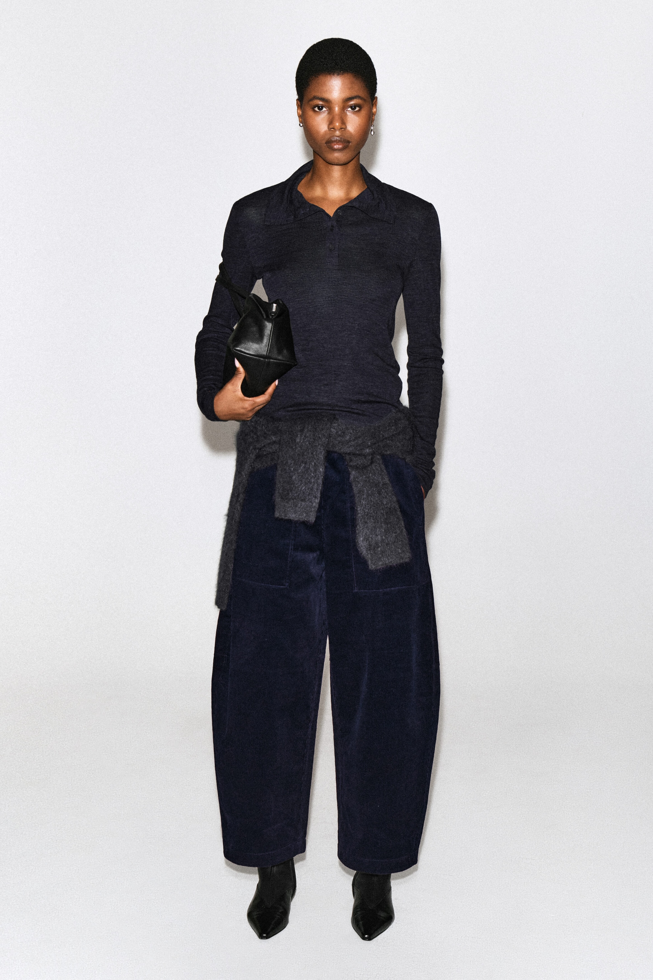 The model is wearing dark navy blue corduroy barrel-leg pants, featuring a wide, curved silhouette and front patch pockets. The pants are styled with a dark long-sleeve polo shirt, a grey knit sweater tied around the waist, and black pointed-toe boots. The model is wearing dark navy blue corduroy barrel-leg pants, featuring a wide, curved silhouette and front patch pockets. The pants are styled with a dark long-sleeve polo shirt, a grey knit sweater tied around the waist, and black pointed-toe boots.