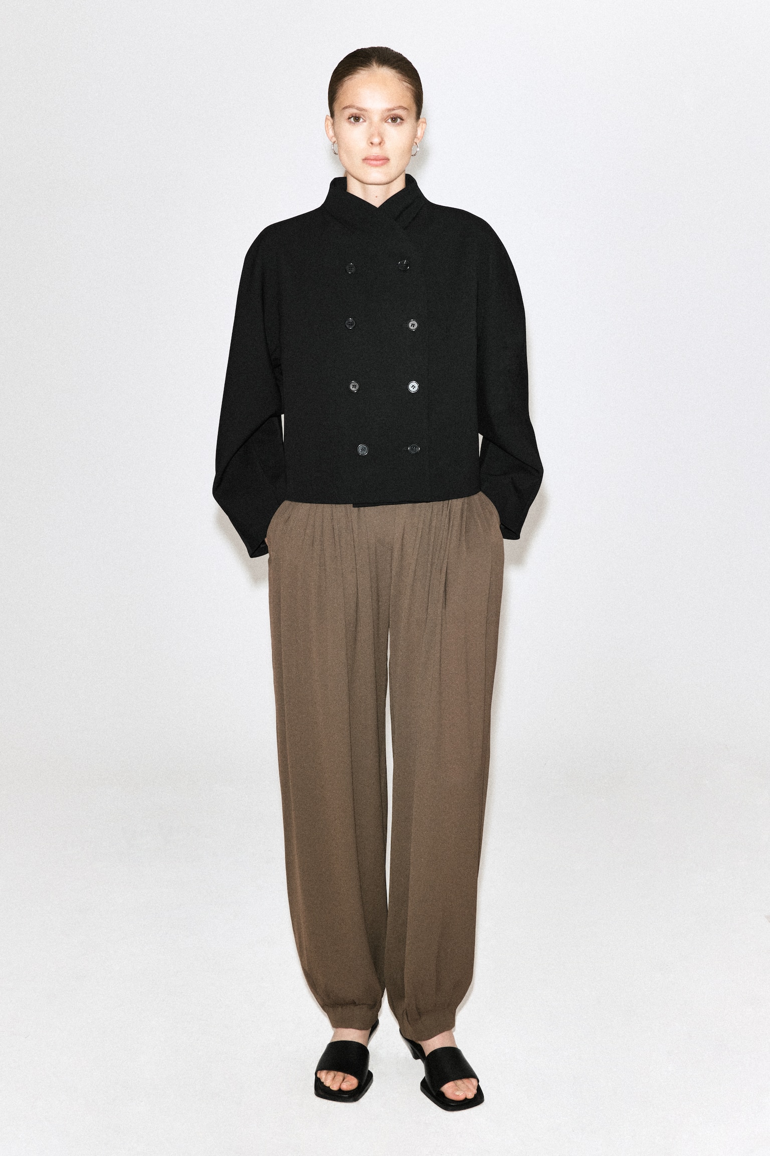 Unos pantalones globo de crepé plisado en tono marrón los lleva la modelo, con una silueta relajada, pliegues en la cintura y bajos fruncidos en los tobillos. Los pantalones se combinan con una chaqueta negra de doble botonadura y sandalias negras abiertas tipo slide. - 1